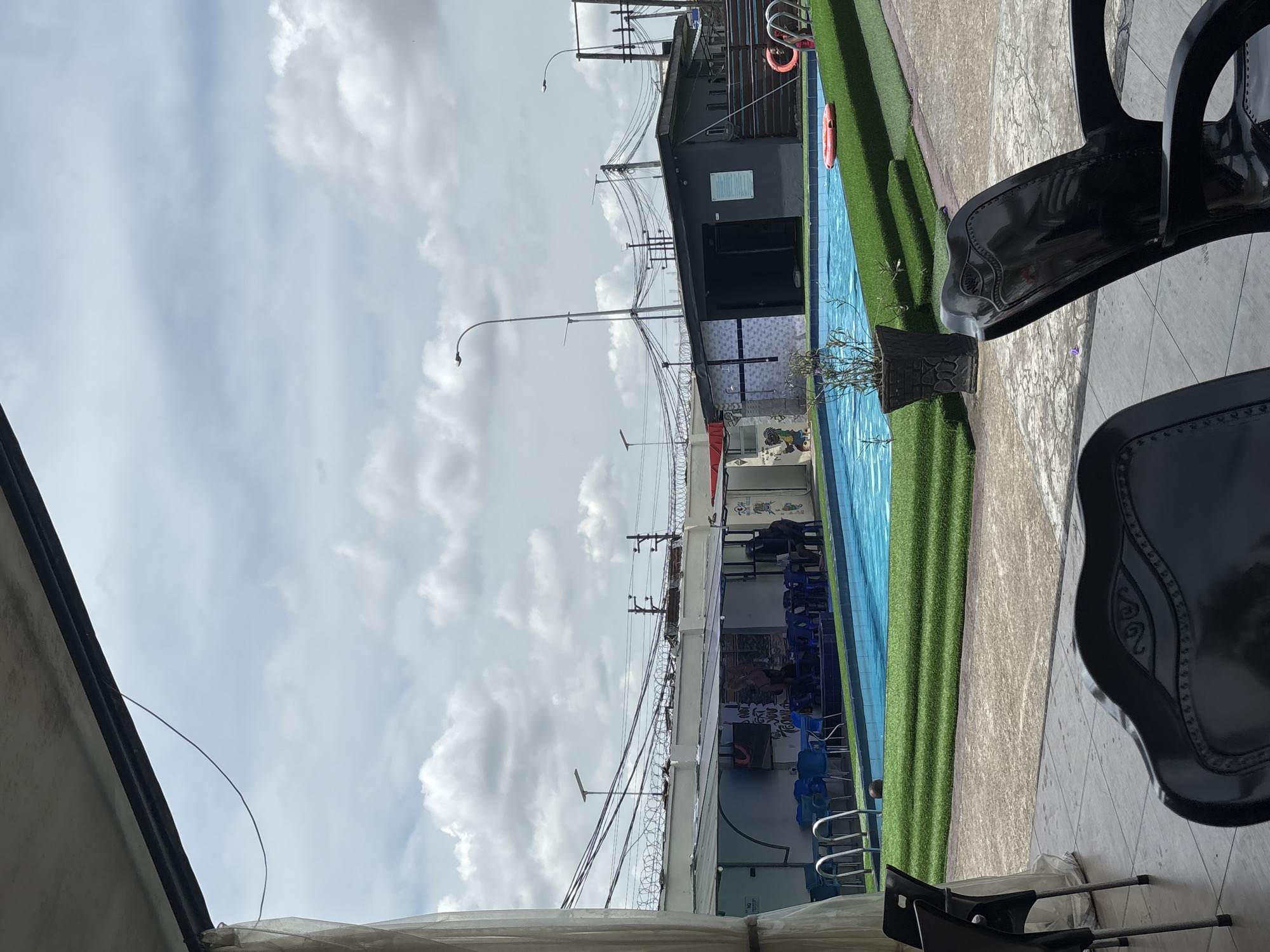 The image shows an outdoor swimming pool area in what appears to be an urban setting, likely in Port Harcourt, Nigeria. The pool has blue water, and its perimeter is lined with artificial green turf. Along one side of the pool, there is a structure with blue chairs and a fence topped with barbed wire. Utility poles and wires are visible above the structure. A life preserver is mounted on the wall.
The sky is cloudy. In the foreground, there are black chairs. The photograph is taken from a low angle, and the camera is positioned close to the chairs. The composition is oriented vertically.