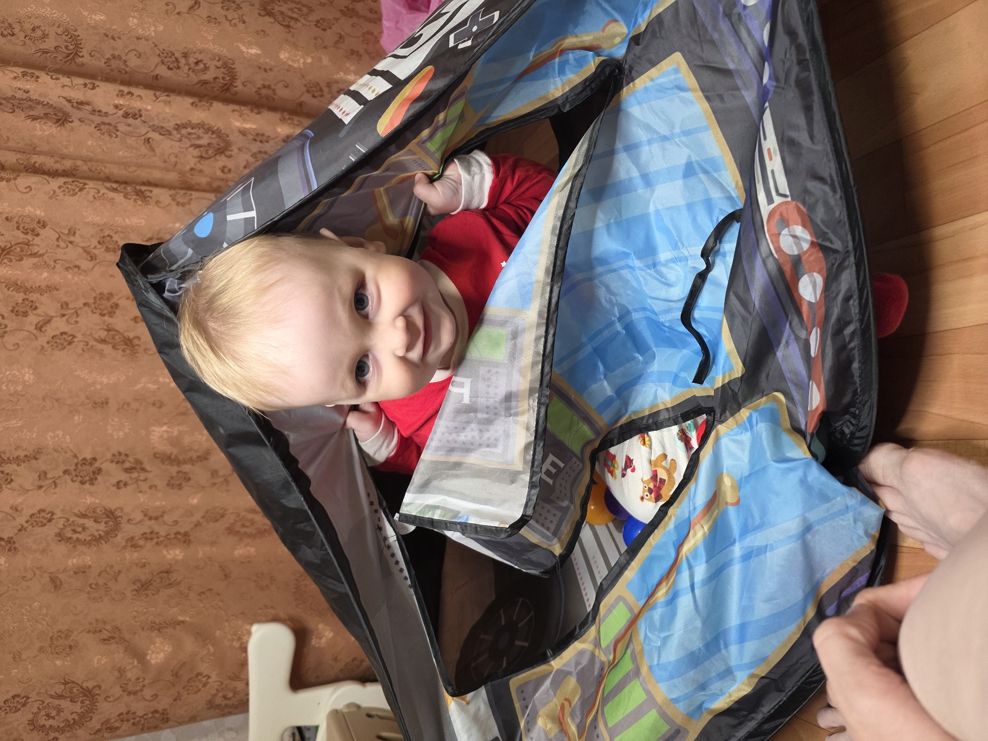 A baby in a red shirt is inside a child's play tent with a blue and gray design. The tent is partially open, with one side held by a fair-skinned adult hand. The background includes a brown patterned curtain and a wooden floor. The location is implied to be Chaikovsky, Russia, but this cannot be independently verified from the image.