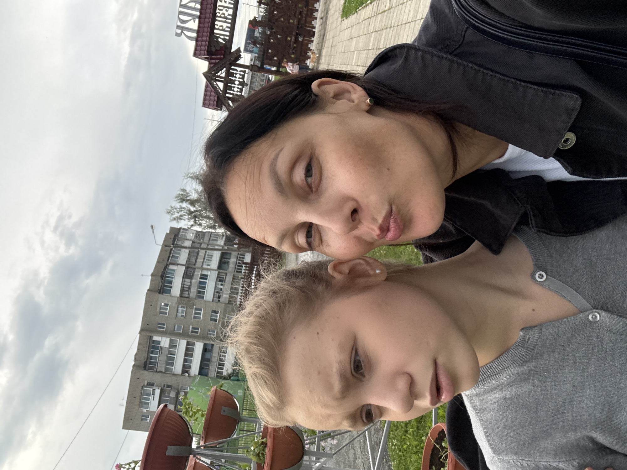 In a selfie taken in Chusovoy, Russia, two females are positioned in the foreground. The female on the left, a younger individual with blonde hair pulled back, wears a gray cardigan. The female on the right, with dark hair, leans in towards the camera with her lips puckered, wearing a dark jacket over a white shirt. The background reveals a brick building, a decorative wooden archway with Cyrillic lettering, and a paved walkway with grassy areas. The sky is overcast.