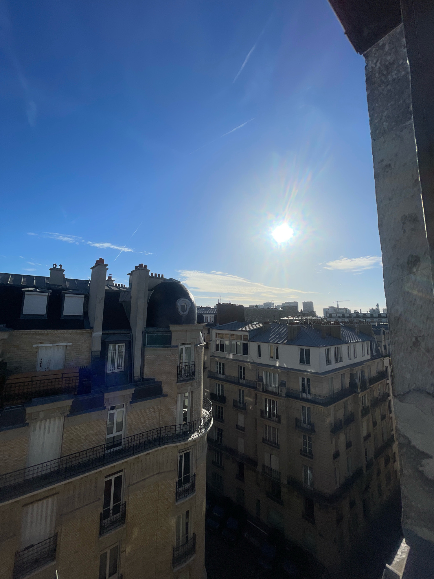The image shows a view of Parisian rooftops and buildings under a bright, sunny sky with scattered clouds. The sun is shining intensely, creating a lens flare. A tall, stone-like structure is visible in the foreground on the right, partially obscuring the view. In the middle ground, we see several apartment buildings with distinctive mansard roofs and dormer windows, typical of Parisian architecture. One building has a rounded, domed section. Chimneys are visible on top of the roofs. The buildings are constructed with brick and stone facades. The sky is a clear blue with thin, wispy clouds and faint contrails from an aircraft. The overall scene depicts a sunny day in a dense urban environment.