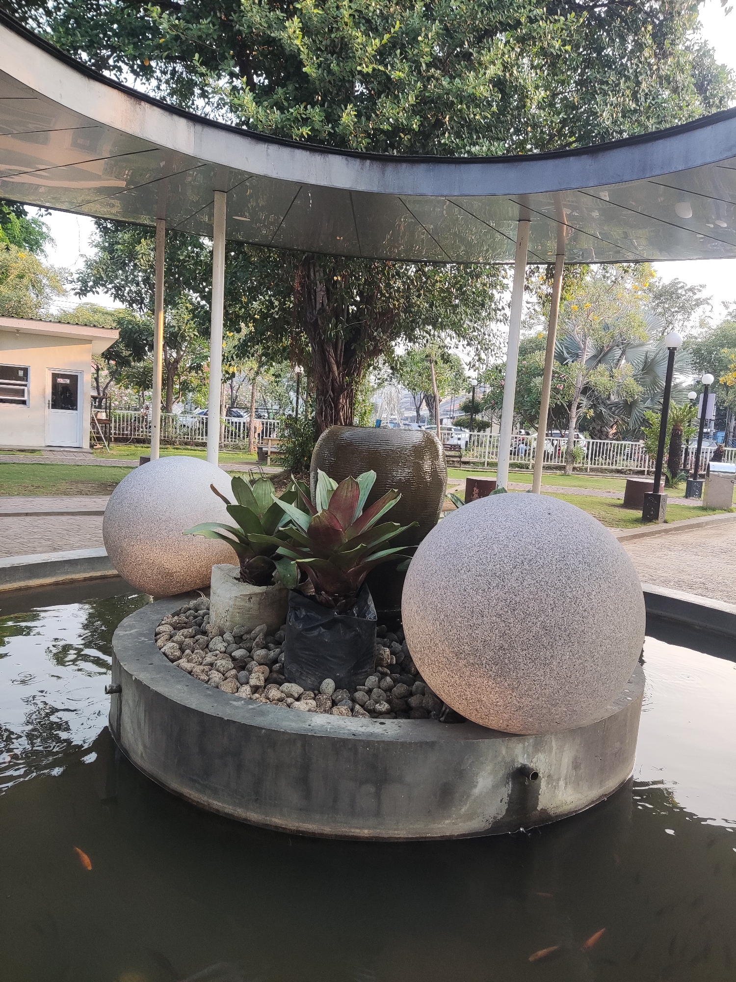 The image shows an outdoor fountain in the city of Pagerwojo, Indonesia. The fountain features a circular concrete base surrounded by water, with three large decorative spheres and several plants. Two of the spheres are made of speckled gray stone, and one is a dark, rounded vase-like object, possibly a water feature. The plants, including bromeliads, are in pots within the fountain's central structure. The water contains several small, orange fish. In the background, a building with white walls and a blue roof is visible, along with trees, a paved walkway, and a white fence. The overall setting appears to be a park or public garden, with a large tree canopy overhead.