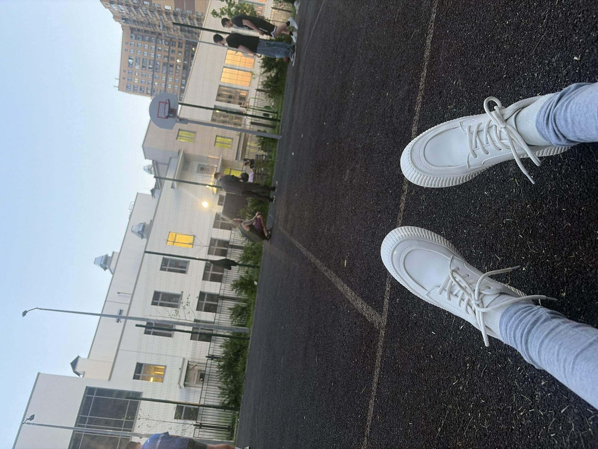 The image shows a low-angle shot of a person standing on a basketball court. The focus is on their white sneakers and gray leggings. In the background, there's a basketball hoop and several people, some appearing to be playing basketball or hanging around the court. Buildings are visible in the backdrop, including a high-rise apartment building and a lower, white building with multiple windows. The lighting suggests it is dusk or early evening. The court surface is black asphalt.
Without additional information, it is impossible to verify that this image was taken in Saint Petersburg, Russia.