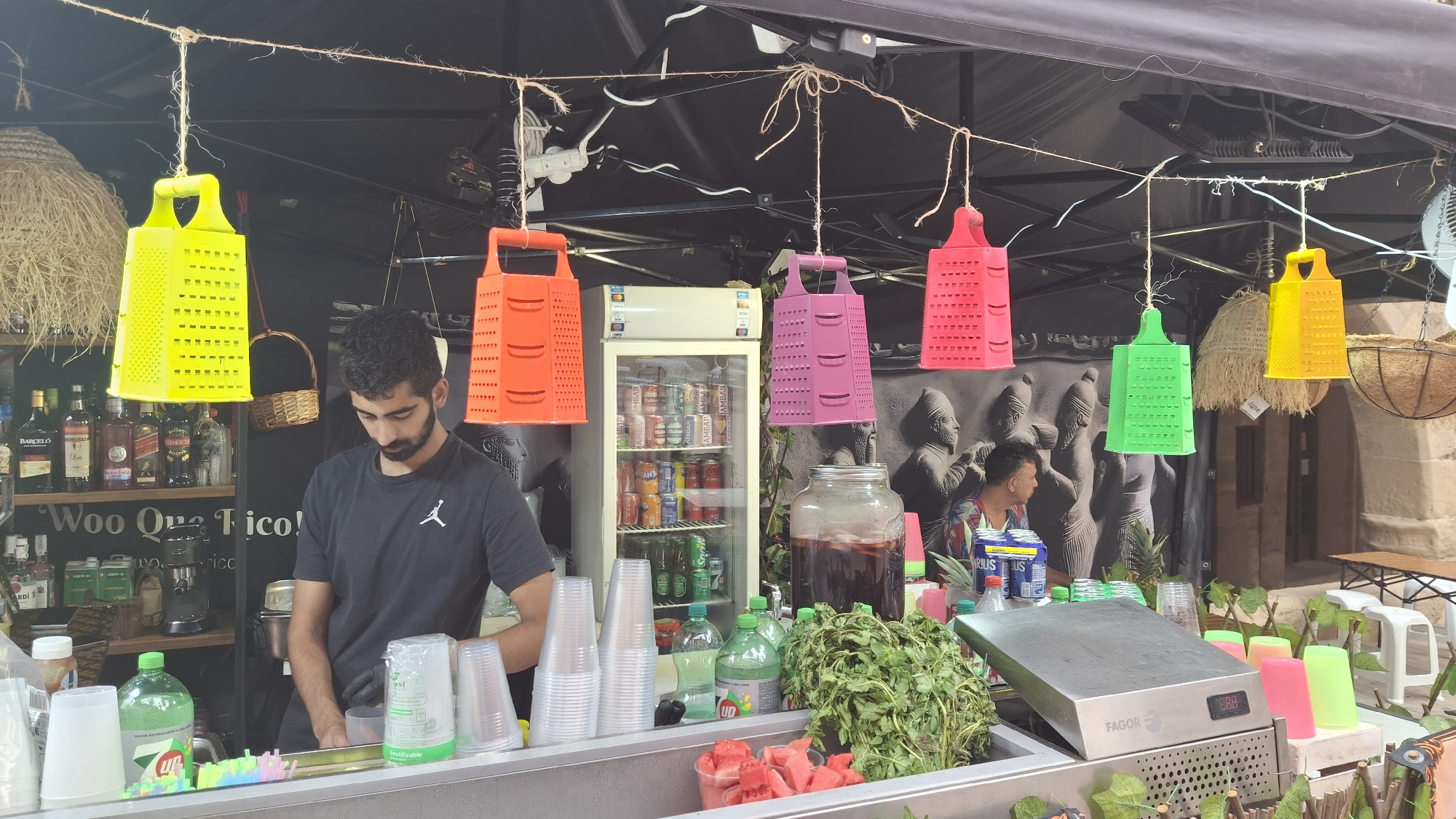 In La Fresneda / la Freixneda, Spain, a man with dark, curly hair and a beard, wearing a black t-shirt with a white logo, is working behind a counter at what appears to be an outdoor food or drink stand. He is looking down, possibly preparing something. In front of him on the counter are stacks of clear plastic cups, some with colorful straws, and a green bottle. Behind him, shelves are stocked with various bottles of liquor and other beverages. A refrigerator filled with canned drinks is visible to his right. Suspended from a string above the counter are several brightly colored, grater-shaped decorations in yellow, orange, pink, and green. To the right of the refrigerator, a large glass jar contains a dark liquid, likely a beverage. Further to the right, another person, partially visible, is seated, looking to the left. The background suggests an outdoor setting with stone architecture and some tables and stools. The overall scene depicts a vibrant market or street food stall environment.