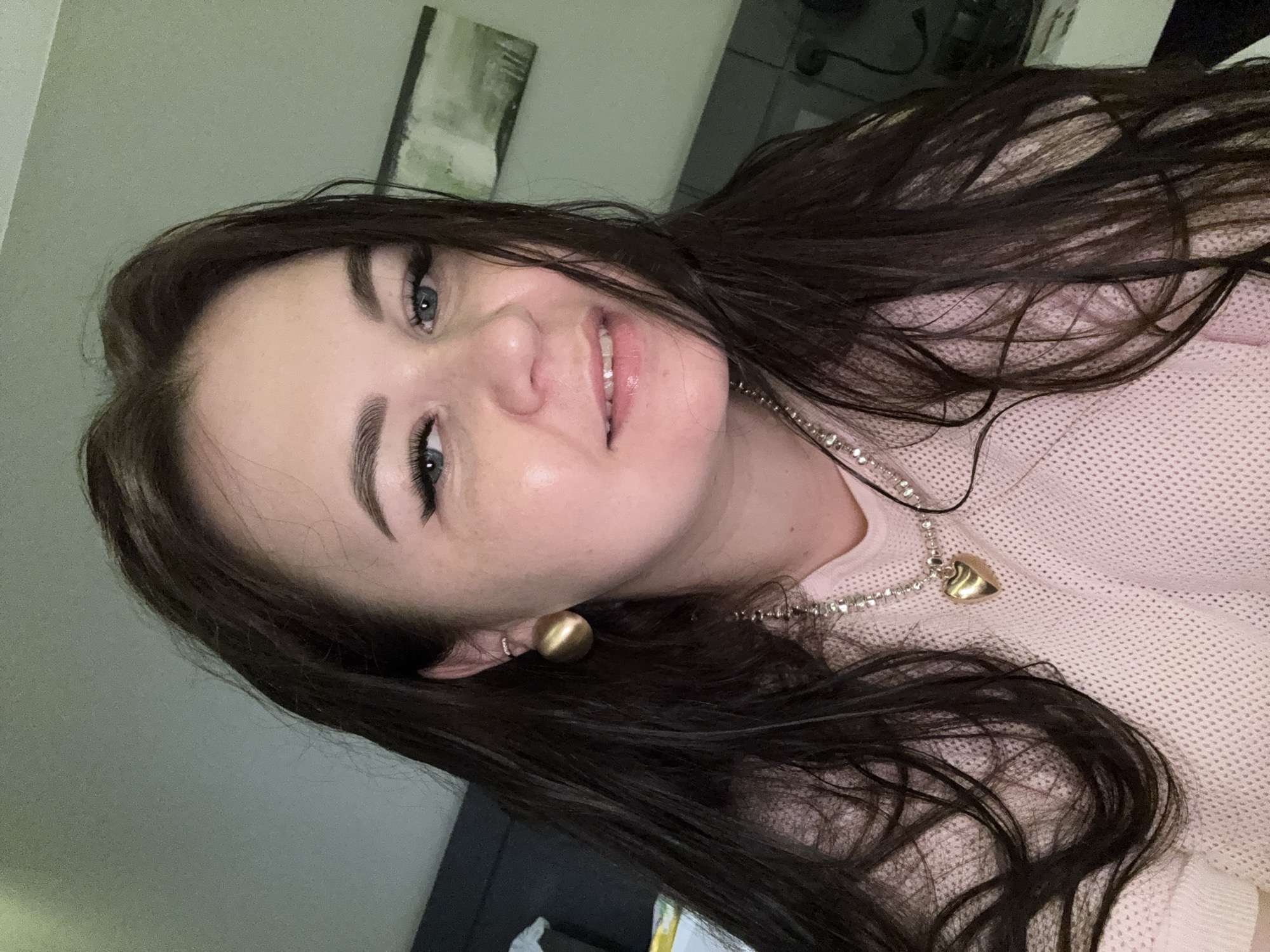 A woman with long, dark brown hair is shown in a close-up, selfie-style portrait. She has light skin with some freckles and is wearing a light pink, mesh-like top. Around her neck, she wears a silver necklace with a large, gold heart pendant and a sparkly embellishment around the neckline of her top. She also has large, gold, spherical earrings. Her makeup includes winged eyeliner and mascara. The background appears to be an indoor setting, with a light green wall and a framed piece of art visible. The image does not provide any information about the city of Alanya, Turkey.