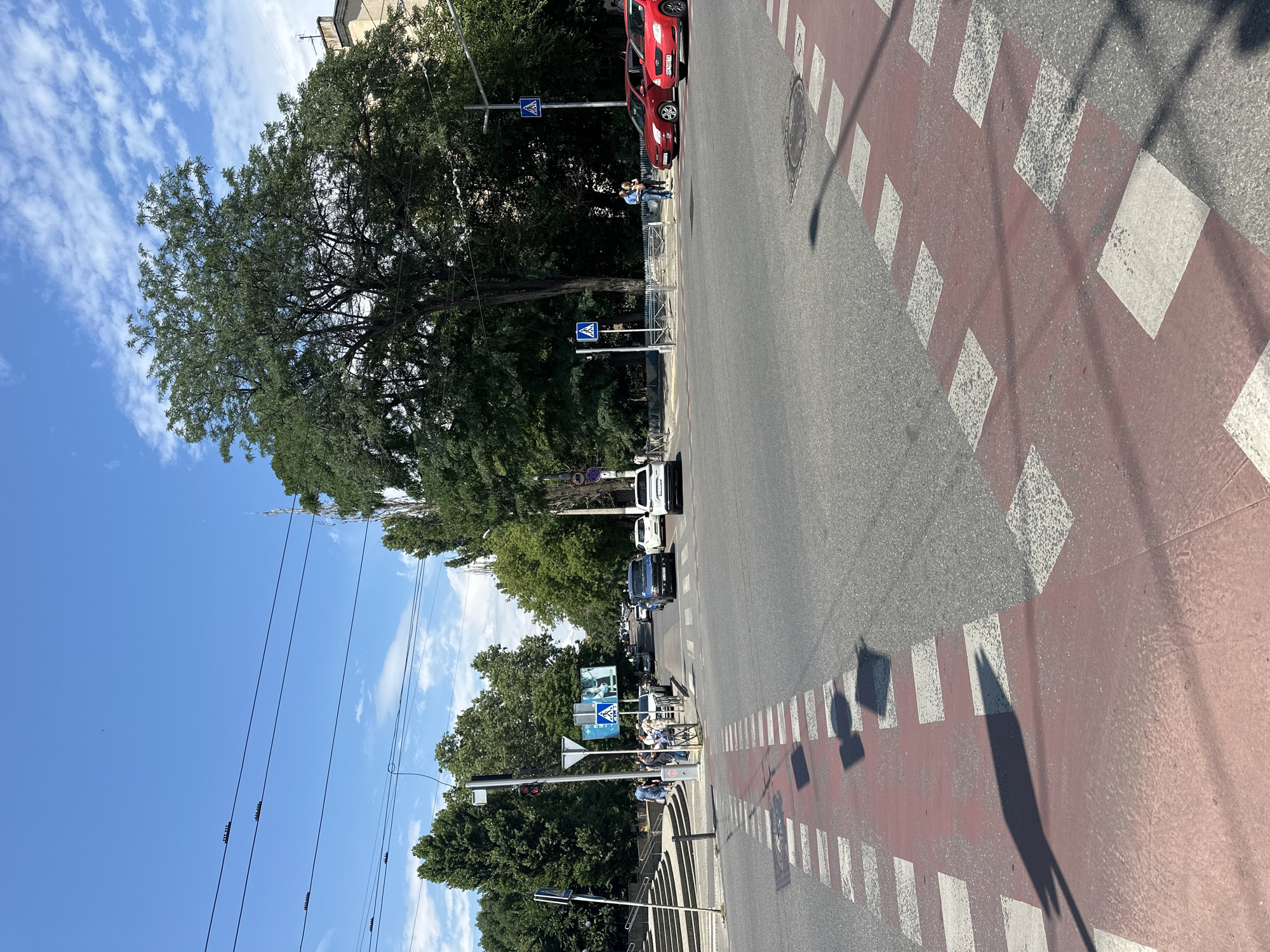 The image depicts a daytime scene at an intersection in Odesa, Ukraine, characterized by a wide asphalt road with clear road markings, including a prominent red and white striped pedestrian crossing in the foreground. Several vehicles are present: a white SUV, a dark-colored van, and two red sedans. A pedestrian crossing sign is visible on the right side of the road. In the background, lush green trees line the streets, and a building with architectural details can be seen. The sky is blue with scattered white clouds. Several traffic lights and utility poles are also integrated into the urban landscape. A group of people are gathered near the pedestrian crossing, suggesting active city life. The overall impression is of a busy city intersection on a clear, sunny day.