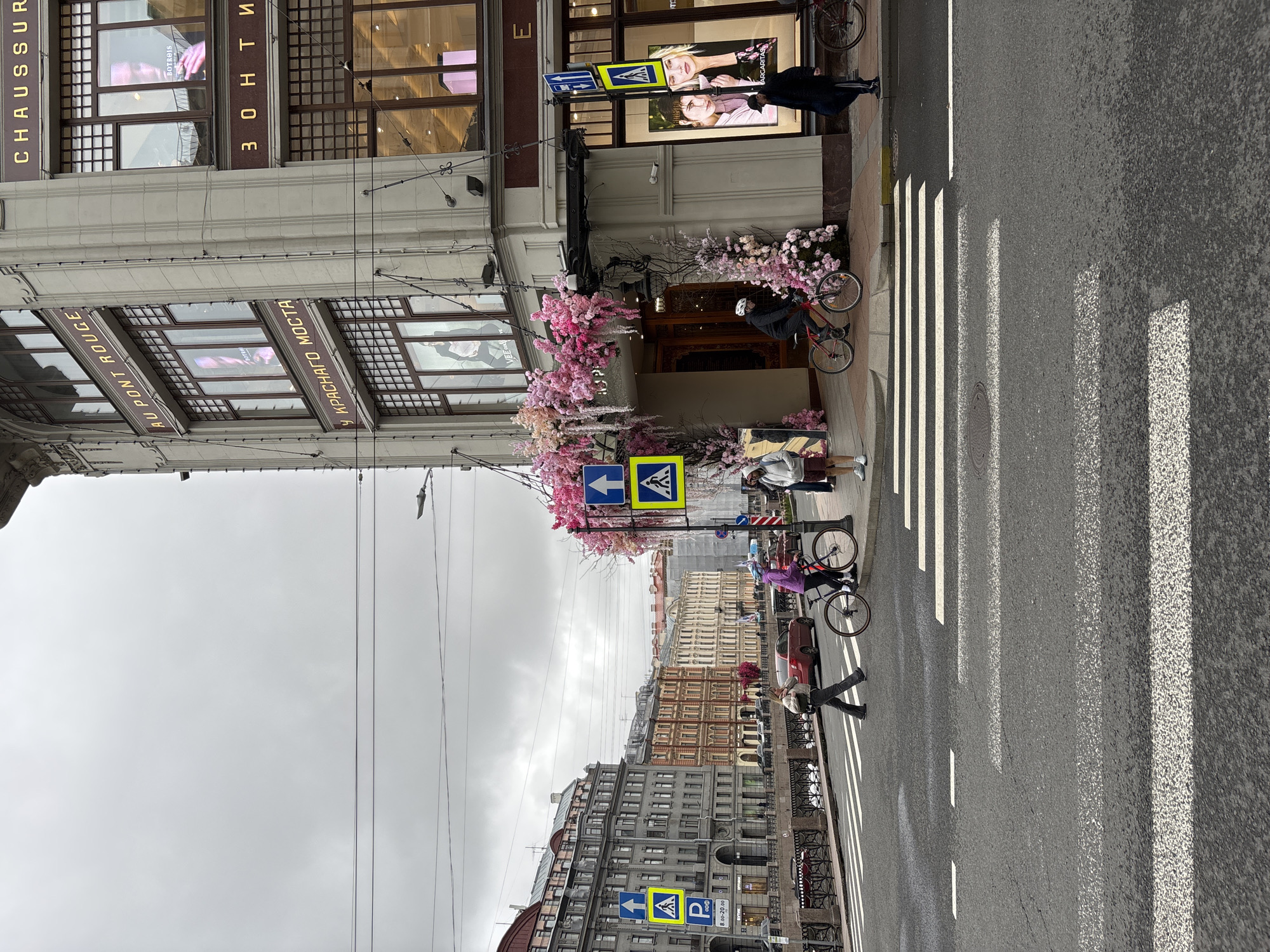 The image captures a street scene in Saint Petersburg, Russia, oriented vertically. The sky is overcast. The foreground features a pedestrian crossing with zebra stripes. On the right, a portion of a building is visible. On the ground floor, there is a doorway decorated with pink floral arrangements, and people are passing by, including a person on a bicycle. Above the doorway, a sign with a pedestrian symbol is visible. The building’s façade features signs with the words "CHAUSSUR" and "ЗОНТИ" written vertically, and "AU PONT ROUGE" and "У КРАСНАГО МОСТА" horizontally. The image also captures other buildings across the street, with architectural details and a similar overcast sky above. Wires are strung overhead, adding to the urban scene. People are walking across the crosswalk, including one with flowers, and there is a car parked on the street. Another pedestrian crossing sign is visible further down the street.