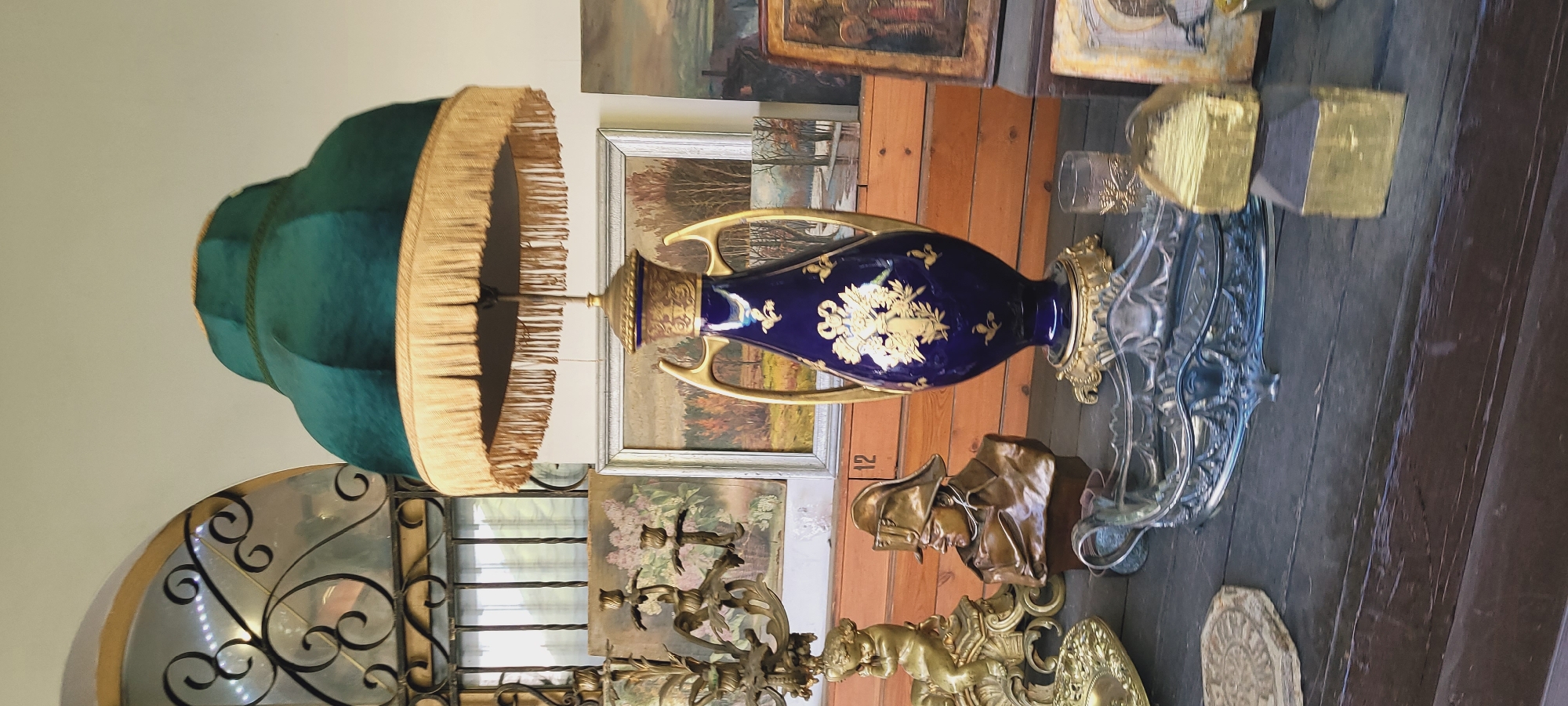 The image shows a dimly lit indoor scene cluttered with various objects. A blue, vase-shaped lamp with gold handles and floral design is centrally located. The lamp is topped with a green shade trimmed with gold fringe. Behind the lamp are several framed paintings leaning against a wooden wall. Other items in the scene include a bronze bust, a silver decorative object, and a candelabra. The scene appears to be an antique shop or a storage area. The city and region are unknown.