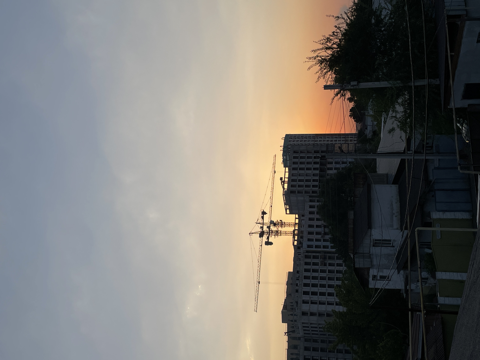The image depicts a twilight scene in the city of Almaty, Kazakhstan, featuring a construction site with cranes and unfinished buildings silhouetted against a gradient sky of soft orange and pale blue. In the foreground, a cluster of low-rise residential structures with weathered facades and corrugated roofs are visible, interspersed with trees and utility poles carrying a network of electrical wires. The larger, modern buildings under construction are to the left and center-right, their concrete structures stark against the dusk. The overall impression is one of urban development juxtaposed with older, lived-in areas, under the fading light of day. No individuals are present in the frame.