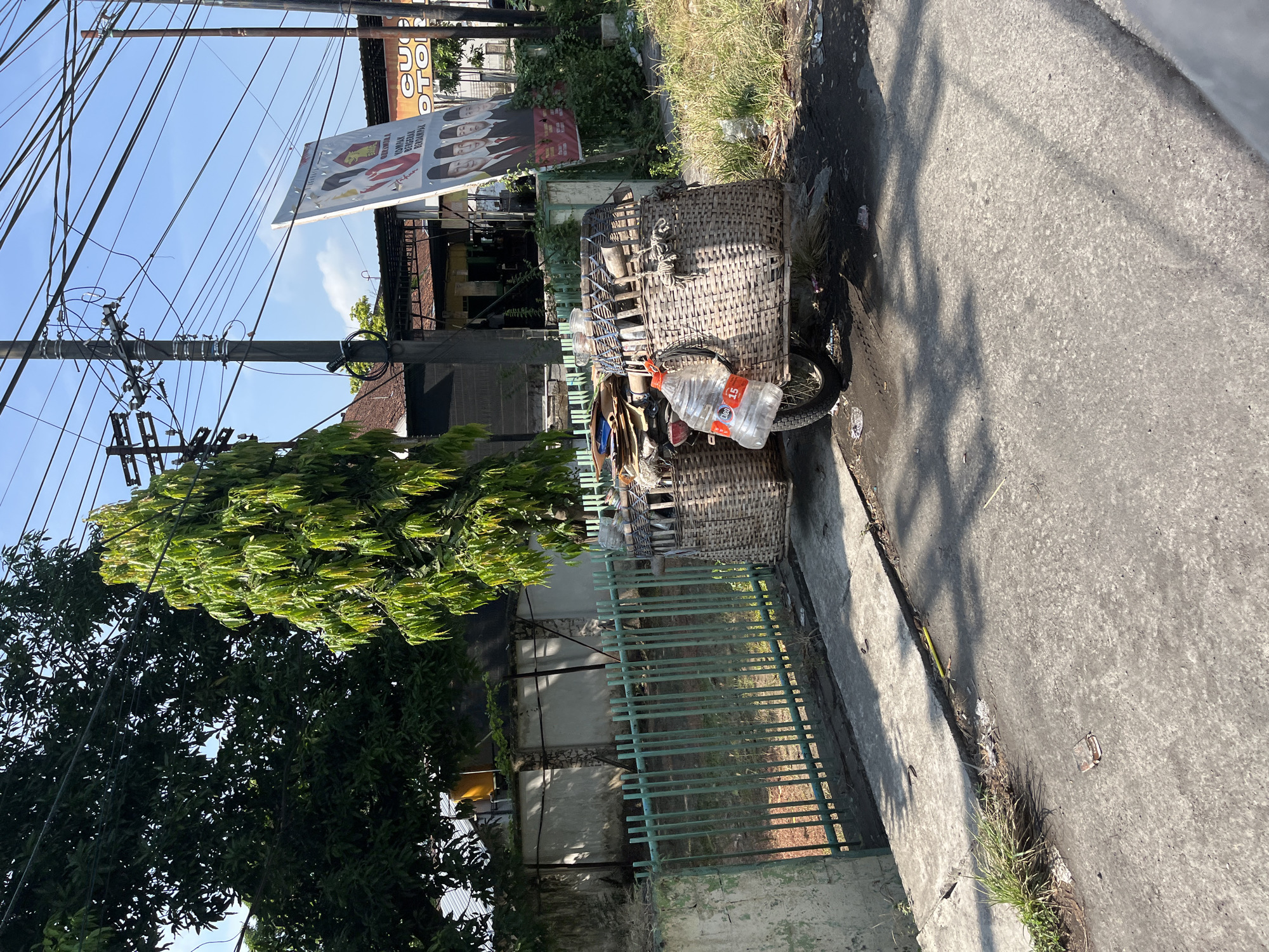 The image depicts a street scene in Tulangan, Indonesia, on a bright, sunny day. A modified motorcycle with a sidecar is parked on the side of the road. The motorcycle is equipped with two large woven baskets on the sidecar, loaded with various items, likely for collection or sale. A prominent feature of the load is a large plastic water bottle, partially filled with a clear liquid and bearing the label "NEO 15".
Behind the motorcycle, a green metal fence runs along the edge of a property. Beyond the fence, there is a low concrete wall and some trees. In the background, a large political billboard featuring several individuals in formal attire is visible, alongside a building with a corrugated metal roof. Numerous power lines crisscross the sky, suspended from utility poles. The ground surface is a mixture of paved road and dirt or gravel, with some scattered debris. The overall impression is one of a typical, perhaps utilitarian, street scene in an Indonesian town.
