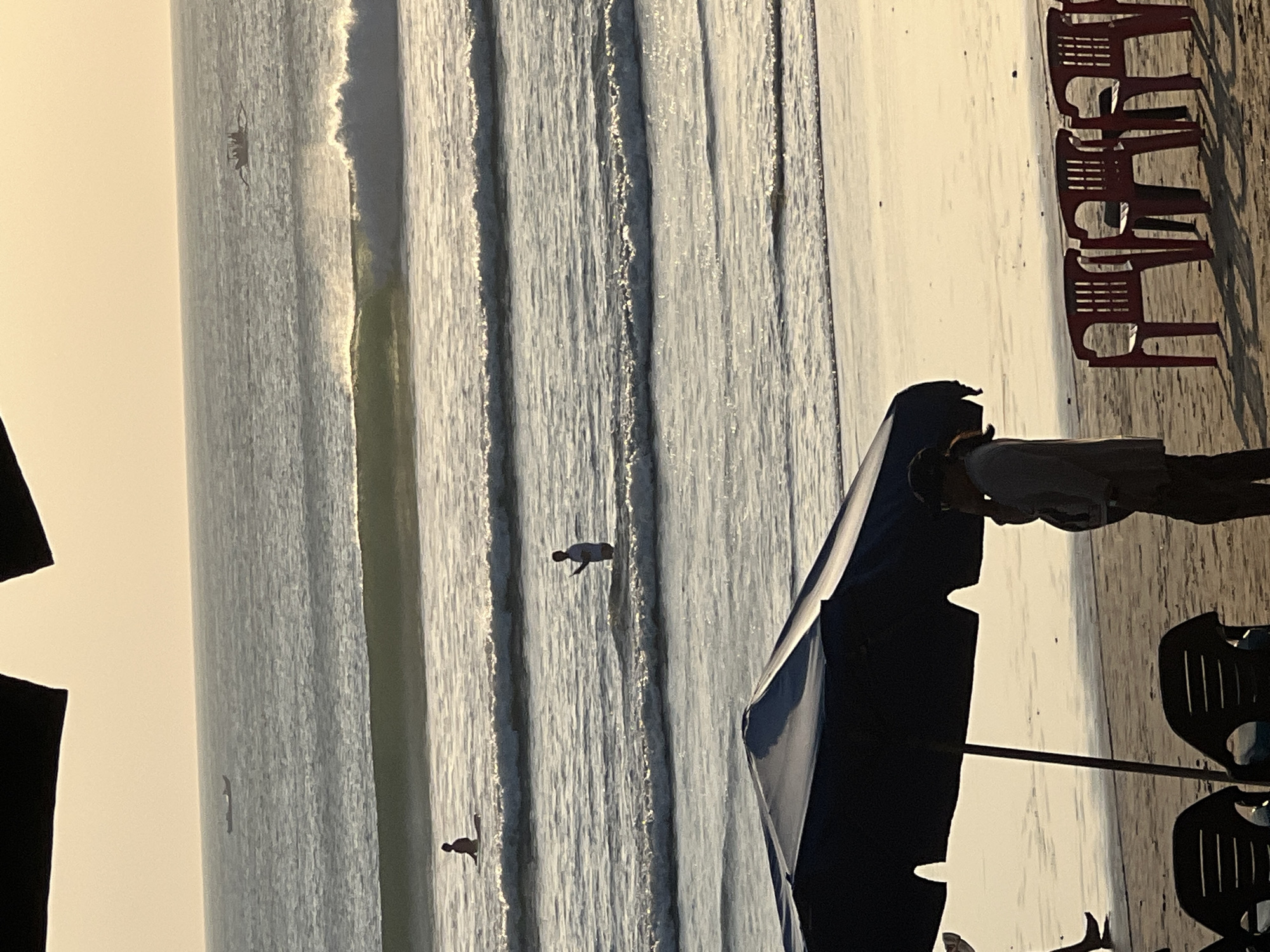 The image depicts a beach scene in Kuta, Indonesia, at sunset. In the foreground, a dark blue beach umbrella casts a shadow on the sand, with two black chairs positioned beneath it. To the right of the umbrella, a person stands facing the ocean, holding a phone to their ear. Further out in the water, another person is standing, and beyond them, a surfer is seated on a surfboard. In the distance, two boats are visible on the ocean, with waves breaking. The sky is a soft yellow, indicating the setting sun, and the ocean is calm with gentle waves. The overall scene is serene and captures the essence of a peaceful evening at the beach.