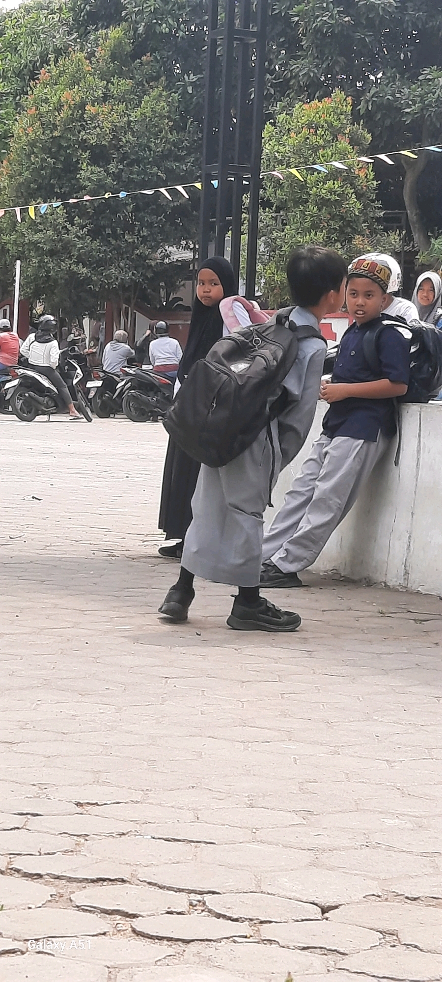 In a sunlit outdoor scene, possibly in Tangkil, Indonesia, a group of children stand on a paved area with a textured surface, suggestive of cobblestones or large pavers. Two boys are engaged in conversation, one leaning against a low, concrete wall, the other standing closer. Both boys wear school uniforms. The boy on the right wears a dark blue short-sleeved shirt, grey trousers, and a patterned skullcap. He is looking towards the boy on the left, who is dressed in a light grey long-sleeved shirt and grey trousers, with a black backpack slung over his shoulder. A young girl in a black hijab and long black dress stands to the left, also wearing a backpack. Her gaze is directed towards the left, away from the boys. In the background, several motorcycles are parked, and people are seated or standing near them, some wearing helmets. Beyond the motorcycles, trees with green foliage form a backdrop, with a black metal fence or structure visible in the upper portion of the image. Small, colorful triangular flags are strung across the scene, adding a festive touch. The overall atmosphere appears casual, perhaps indicating a school break or a gathering.