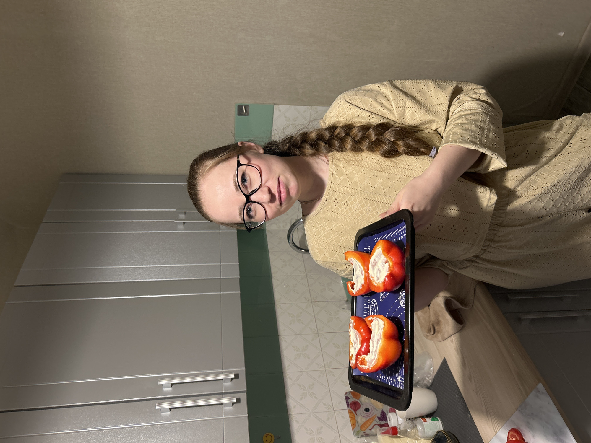 A young adult woman with long, braided brown hair and black-framed glasses is depicted in a kitchen setting, looking directly at the camera with a neutral expression. She is wearing a light-colored (beige or tan) long-sleeved top or dress with an intricate patterned texture.
She holds a dark, rectangular baking tray with a blue silicone mat. On the mat are two large, bright red bell peppers, each cut in half lengthwise and generously stuffed with a white, creamy filling, likely prepared for baking. The blue silicone mat features various white markings, including measurements (e.g., "5 6 7 8 9 10 11", "30 35 40 45") and circular patterns.
The background shows modern kitchen elements. To her left are light grey or silver-colored cabinets with horizontal bar handles. Behind her, a section of the wall features light blue-green tiled backsplash. The floor is covered with light-colored tiles displaying a repeating geometric pattern. A light wood-grained countertop extends from the right, with various kitchen items visible, including bottles and what appears to be an orange cutting board decorated with illustrations of vegetables like carrots and onions. The overall lighting suggests an indoor setting, likely evening or night, illuminated by artificial lights. No specific time of day or weather is discernible.