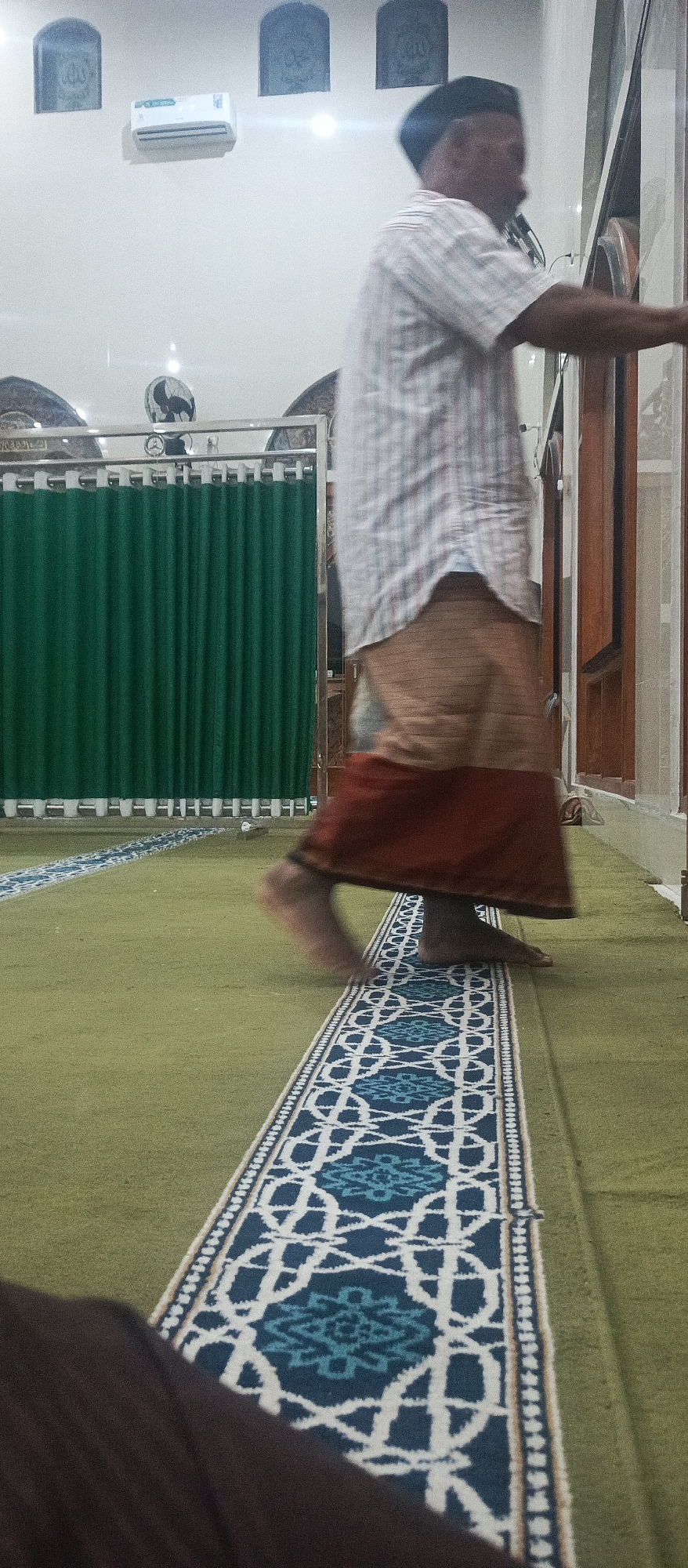 In a room with white walls, an air conditioner is mounted high on the left. Four decorative arched panels are affixed to the wall. A man wearing a checkered shirt, a tan sarong, and a black peci hat is blurred as he walks. His bare feet are visible. A green curtain hangs behind him. The floor is covered with a light green carpet, with a patterned runner featuring white and blue geometric designs. The scene is located in the city of Arjasa, Indonesia.