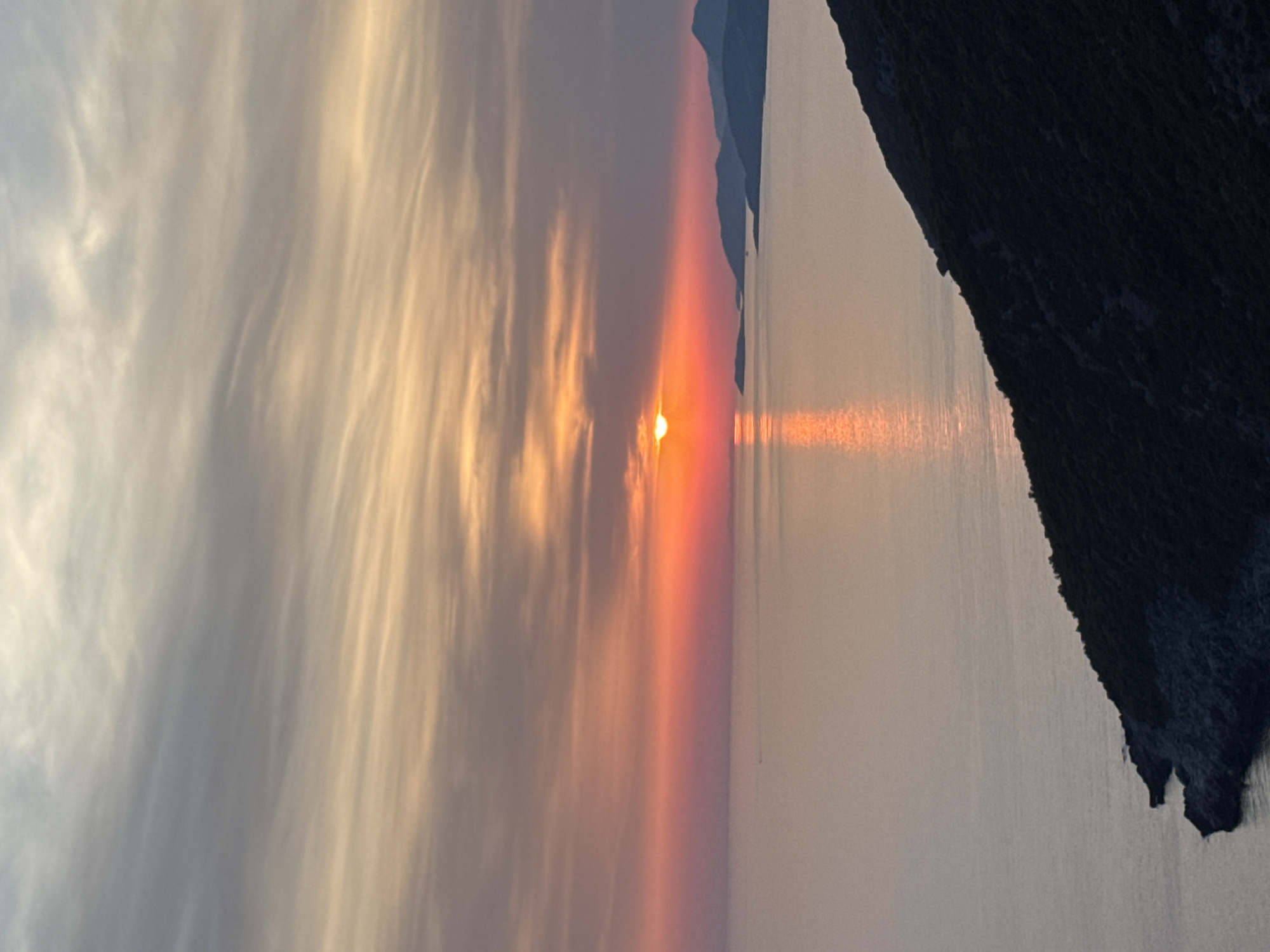 The image shows a sunset over the sea, with a silhouetted mountainous coastline. The sky is filled with streaks of orange, pink, and gray clouds. The sun, a bright orange orb, is low on the horizon, casting a shimmering reflection on the water. In the foreground, a dark, densely vegetated hillside slopes down to the sea. The scene appears to be a natural landscape, possibly a coastal area. The location specified, Zagora, Montenegro, is consistent with a mountainous coastal region. There are no people or explicit actions depicted in the image. The overall mood is serene and picturesque.