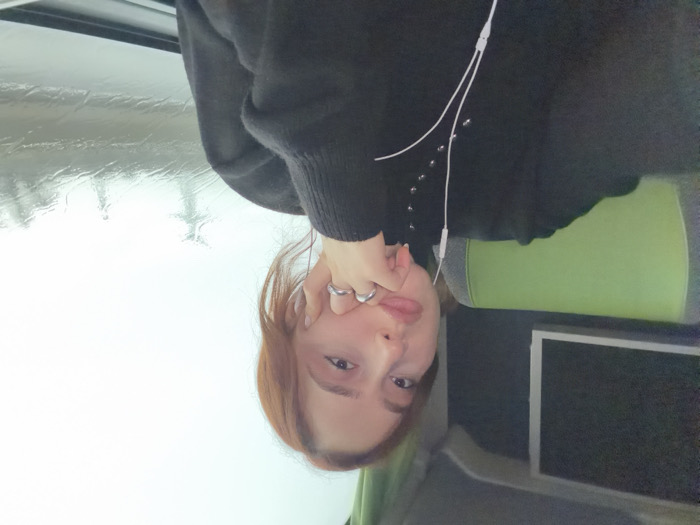 A female individual is seated within a moving vehicle, likely a bus or coach, during daytime overcast or rainy conditions. She is positioned in the foreground, facing the camera directly, capturing a self-portrait. She has reddish-brown hair and fair skin. Her left hand, adorned with a silver-colored ring on the ring finger, is raised with the index finger and thumb supporting her chin. She wears a dark, long-sleeved garment with a high collar and multiple small, metallic studs or buttons arranged vertically on the left chest area. White wired earphones are inserted into her ears, with the wires visible descending along the front of her top. Behind her left shoulder, a distinctive green and grey padded headrest or upper seat portion is visible. To her upper left, a rectangular screen with a dark display is mounted on the vehicle's interior wall. To her right, a window reveals an exterior scene characterized by streaks and droplets of water on the glass, indicative of rain. Through the wet window, indistinct, dark silhouettes, resembling power line towers, are visible against a bright, uniformly overcast sky. The image does not contain specific visual information to independently verify the location as Moineville, France.