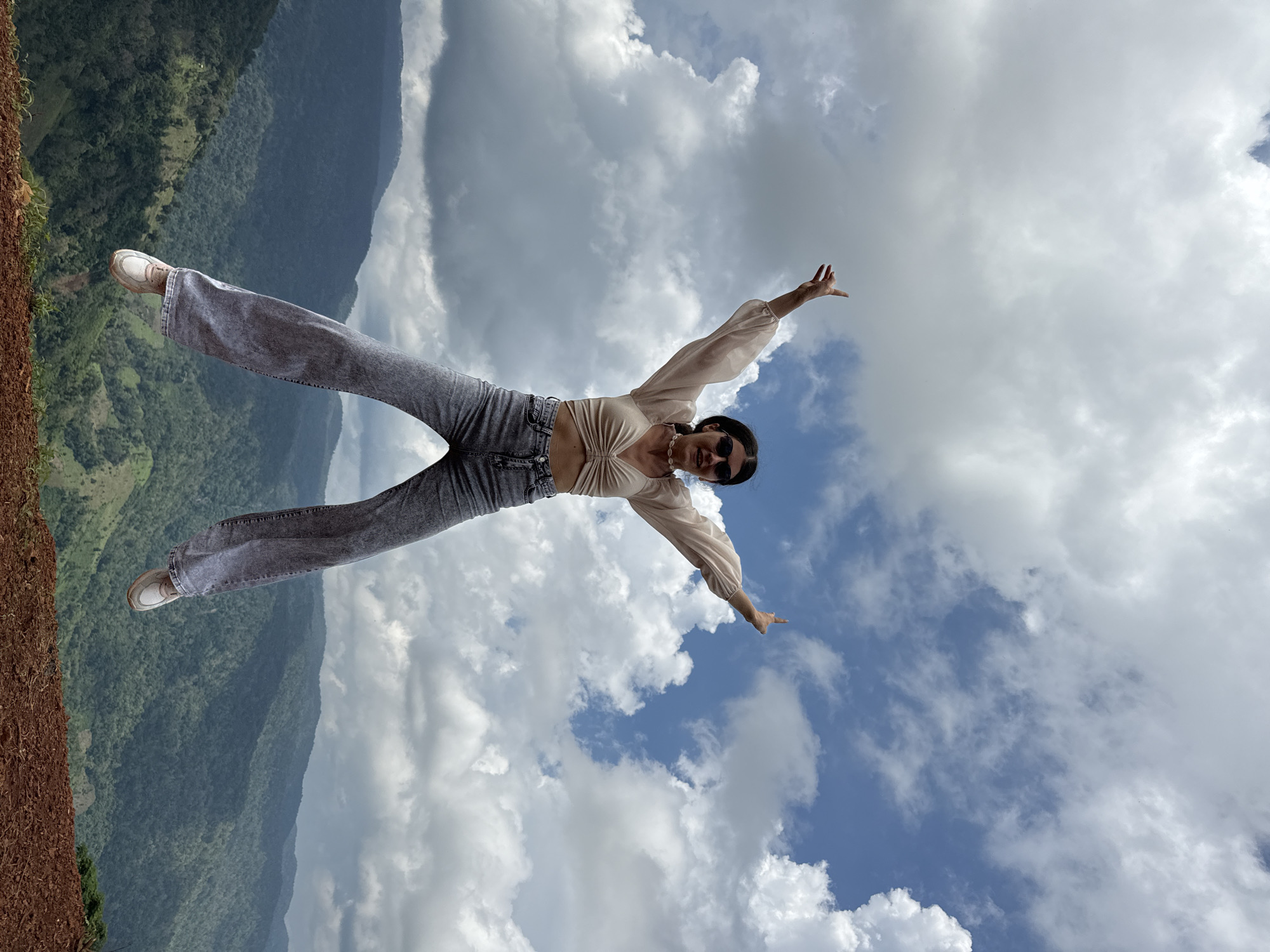 A young woman is captured mid-air, jumping with arms outstretched against a backdrop of a cloudy sky and a lush, mountainous landscape. She is wearing a light-colored, crop-top shirt with billowy sleeves, high-waisted grey flared jeans, and white sneakers. Sunglasses adorn her face, and a pearl necklace is visible. The foreground shows the edge of a dirt path and sparse vegetation. The background is dominated by layers of verdant, mist-covered mountains, characteristic of the highlands. The scene is likely situated in a rural, elevated area such as the mountainous regions of Nao Sẻ, Vietnam, known for its scenic beauty and terraced fields, although no specific urban landmarks are visible. The woman appears to be expressing joy or freedom.