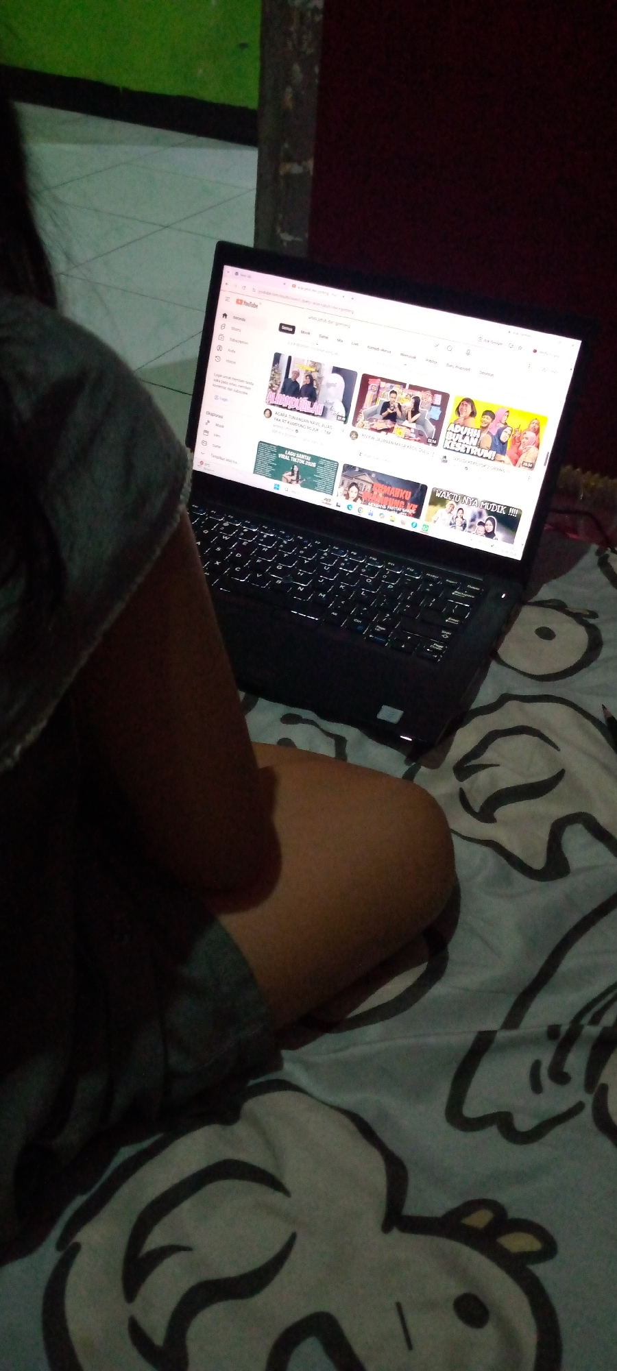 A person is sitting on a bed, facing a laptop which is displaying the YouTube homepage. The person is wearing a grey shirt. The bed has a patterned blanket with what appears to be cartoon characters. The scene is dimly lit, suggesting it is nighttime or the lights are turned off. The background shows a wall and a dark, possibly wooden, surface. The location is identified as Sumberwono, Indonesia.