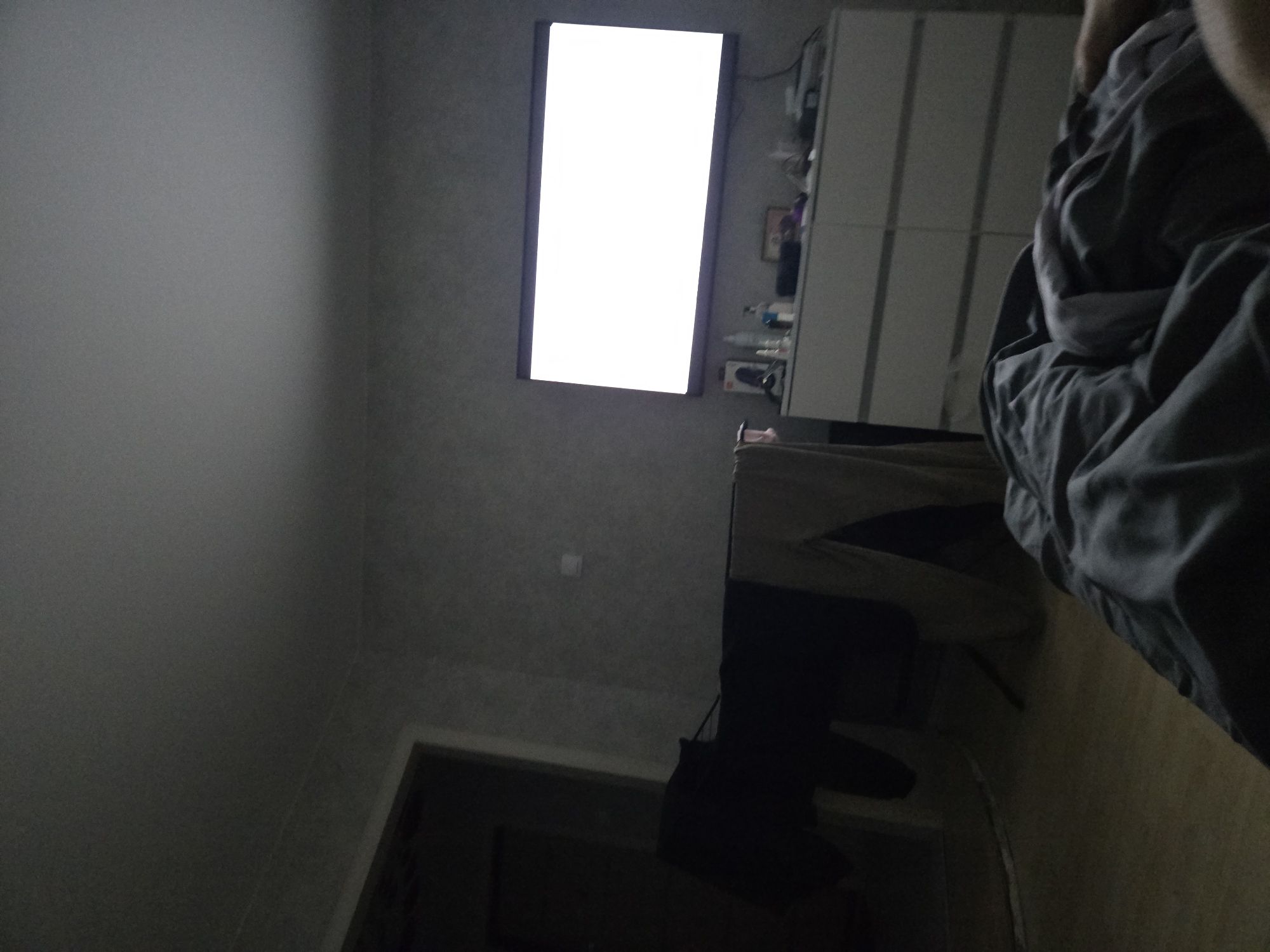 A skewed indoor shot shows a gray-toned room with a white television screen mounted on the wall. To the right, a white cabinet stands beside the screen. A bed with gray blankets is partially visible on the right side of the frame, obscuring some of the cabinet. Below the television, a dark desk and chair are positioned. A partially visible doorway with a dark interior is located at the bottom of the image. Based on the surroundings and the location specified, this appears to be a residential space in Votkinsk, Russia.
