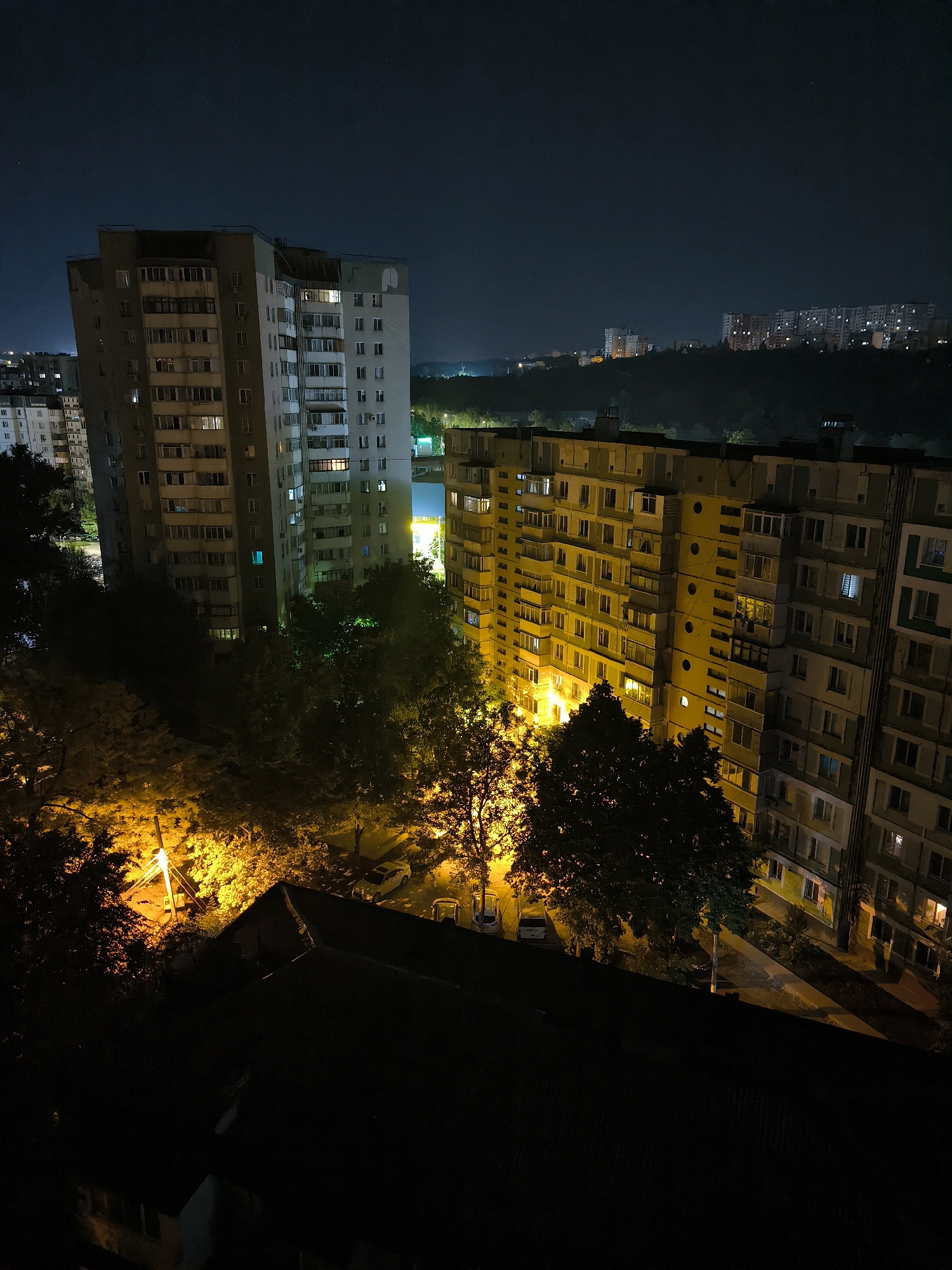 The image depicts a nighttime aerial view of a residential area in Chișinău, Moldova. Two multi-story apartment buildings are prominent in the foreground and midground, illuminated by interior lights and streetlights. The building on the left is a tall, grey and beige structure with numerous windows. The building on the right is a long, yellow building, also with many windows, some of which are lit. Trees are scattered between the buildings, some illuminated by streetlights, casting shadows. The foreground is dominated by the dark roof of another building. In the background, more apartment buildings are visible on a distant hill, faintly lit against the dark sky. The overall scene conveys a sense of urban night life, with the lights from the apartments suggesting occupancy.