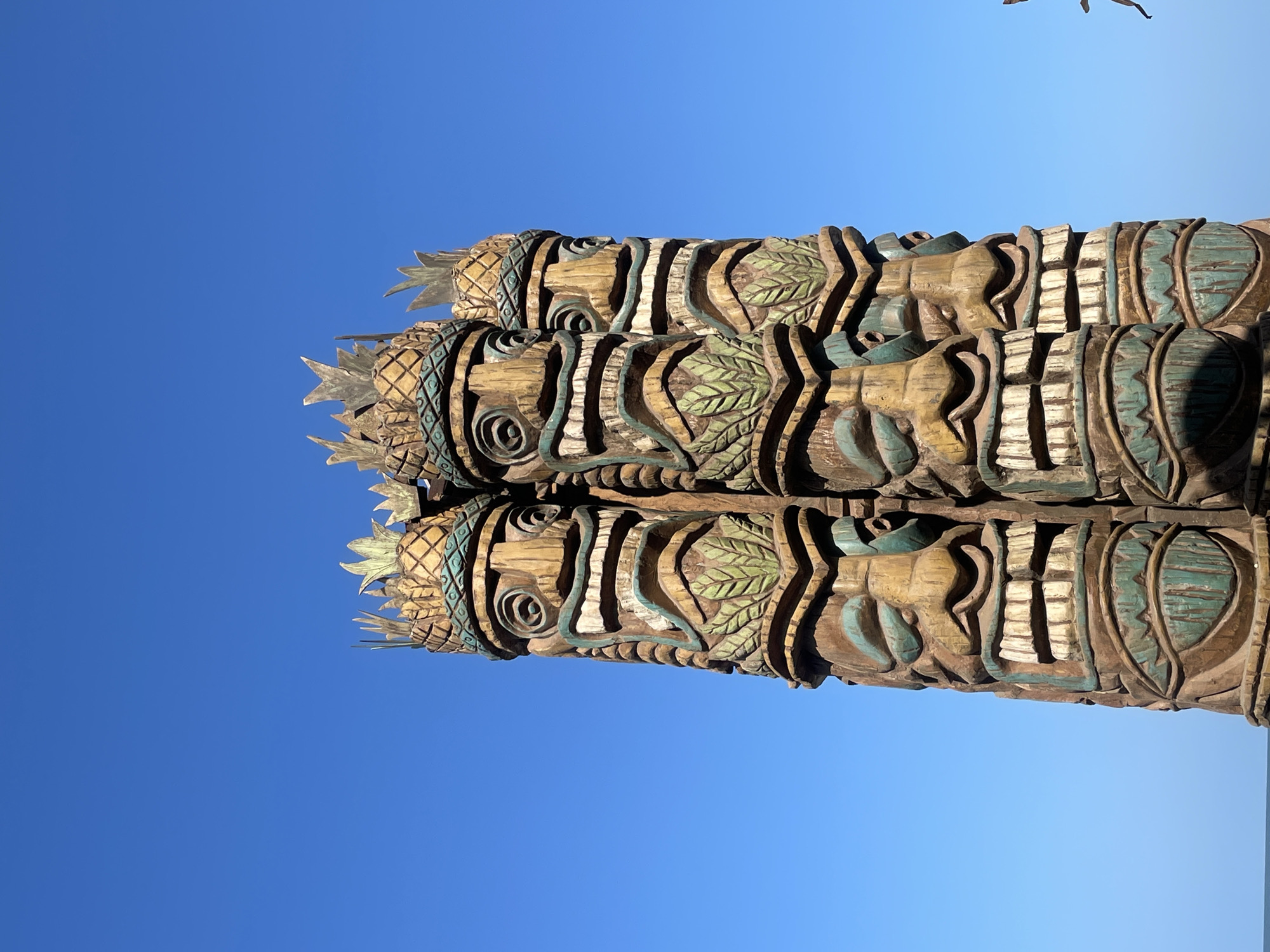 Three carved wooden totem poles stand against a clear blue sky. The carvings include pineapple-shaped tops, faces, leaves, and geometric patterns. The carvings are partially painted with blue and green accents. The location is Chopdem, India.