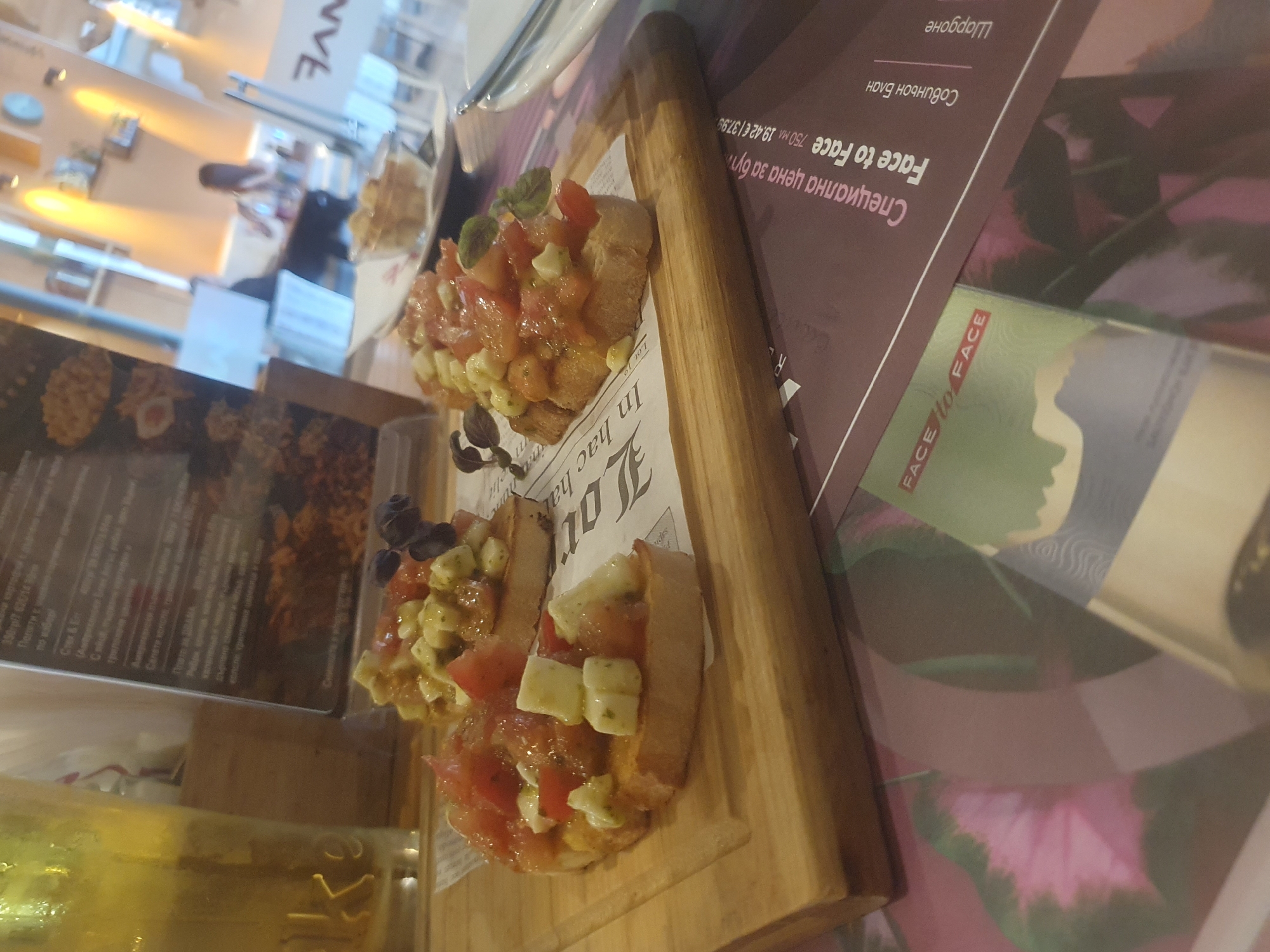 Here's a description of the image:
The image features a close-up shot of bruschetta on a wooden board, next to promotional material for "Face to Face" wine. The bruschetta is topped with tomatoes, basil, and what appears to be mozzarella. The promotional material for the wine is in Bulgarian. The wooden board has printed newspaper-style paper on top of it.
The background suggests a restaurant setting with blurred figures and interior lighting. Based on the OCR text mentioning Bulgarian and the style of food, it is most likely that this scene is located in Plovdiv, Bulgaria.