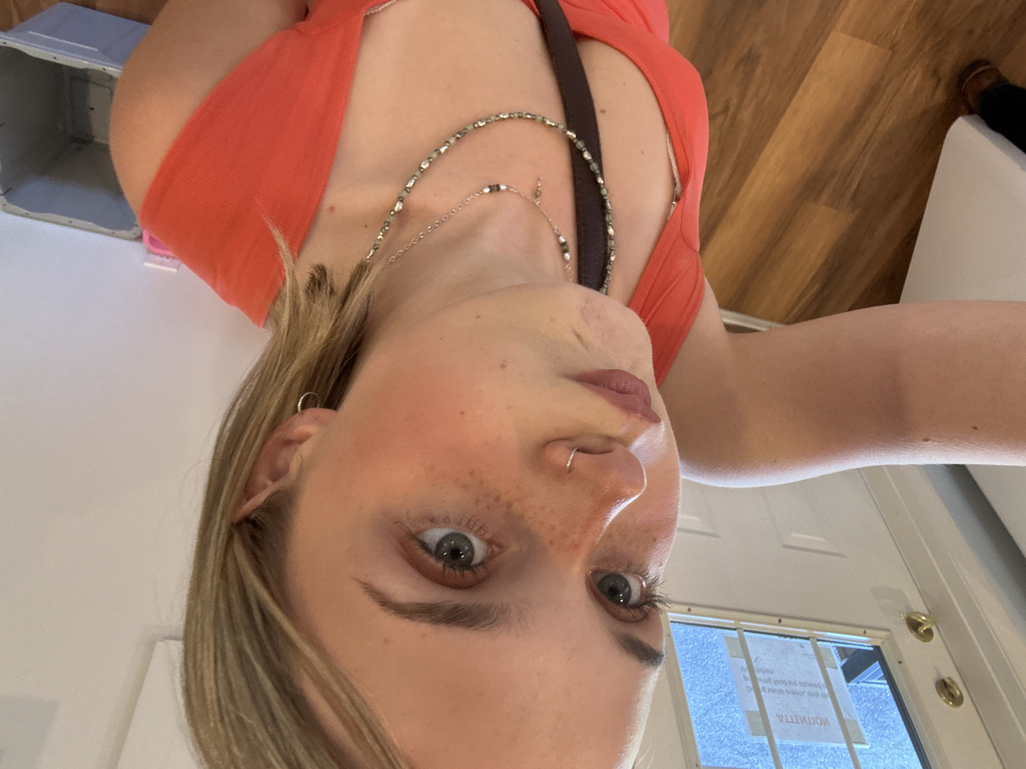 The image is a selfie of a fair-skinned woman with freckles, light brown hair, and grey-green eyes. She is wearing a coral-colored top and a silver necklace, with a dark brown strap visible as well. She has a hoop earring in her left ear and a nose ring in her left nostril.
The background includes a light wood floor, a white door with a gold handle, and a window with a sign. Based on the backward text visible in the window's reflection, the location appears to be on Pandosy Street in Kelowna, Canada.