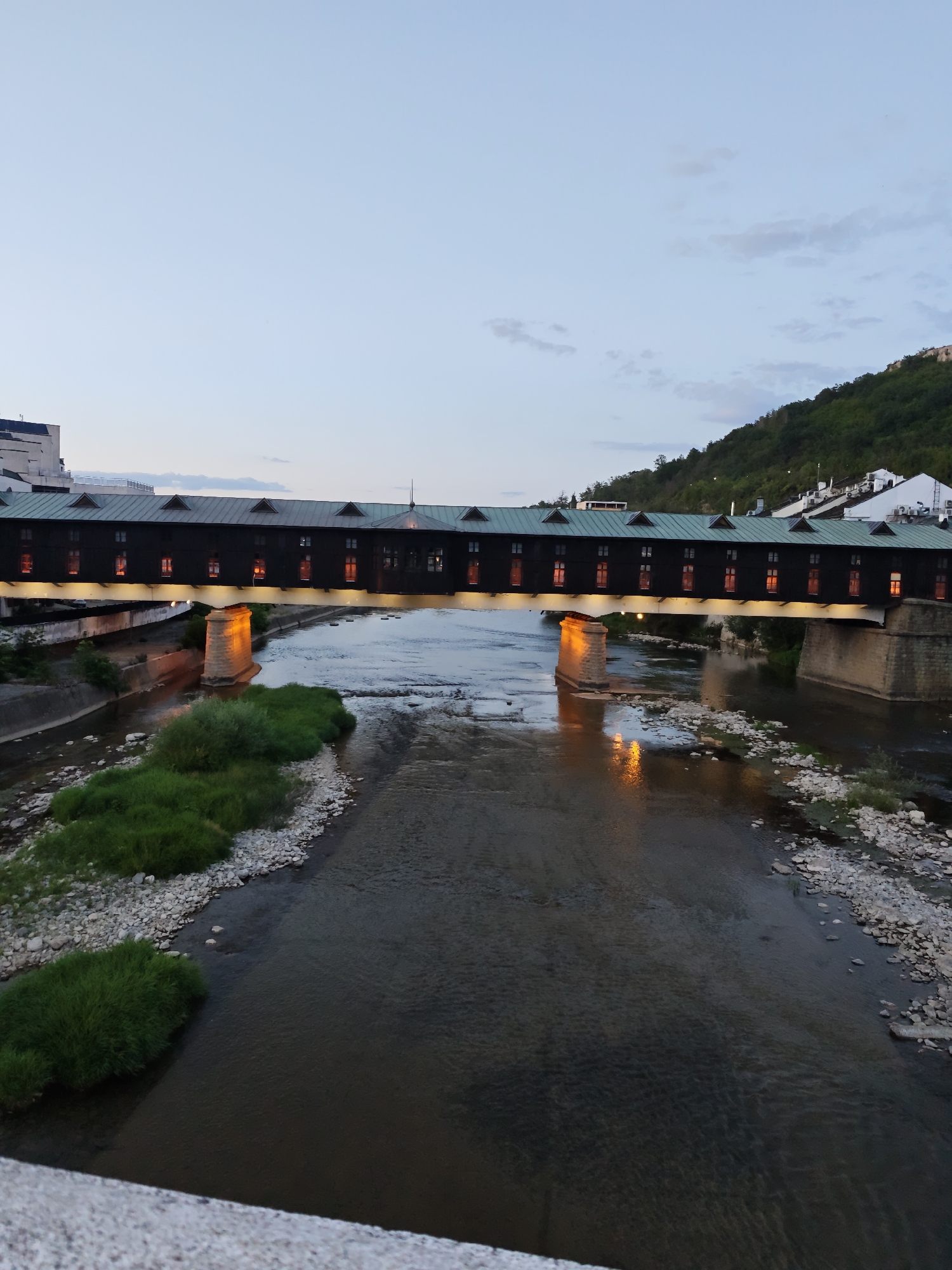 The image captures a twilight view of the Covered Bridge in Lovech, Bulgaria. The long, dark wooden bridge, characterized by its gabled roof and numerous small, illuminated windows, spans a wide, shallow river. The water reflects the warm glow from the bridge's interior lights, creating streaks of orange on the surface.
The riverbed is rocky, with patches of lush green grass growing on the banks and along smaller outcrops within the water. On the right bank, a tree-covered hill rises, with white buildings dotting the slope. The sky is a pale blue, transitioning from day to night, with faint clouds visible. The overall scene is serene and atmospheric, highlighting the architectural and natural beauty of the location.