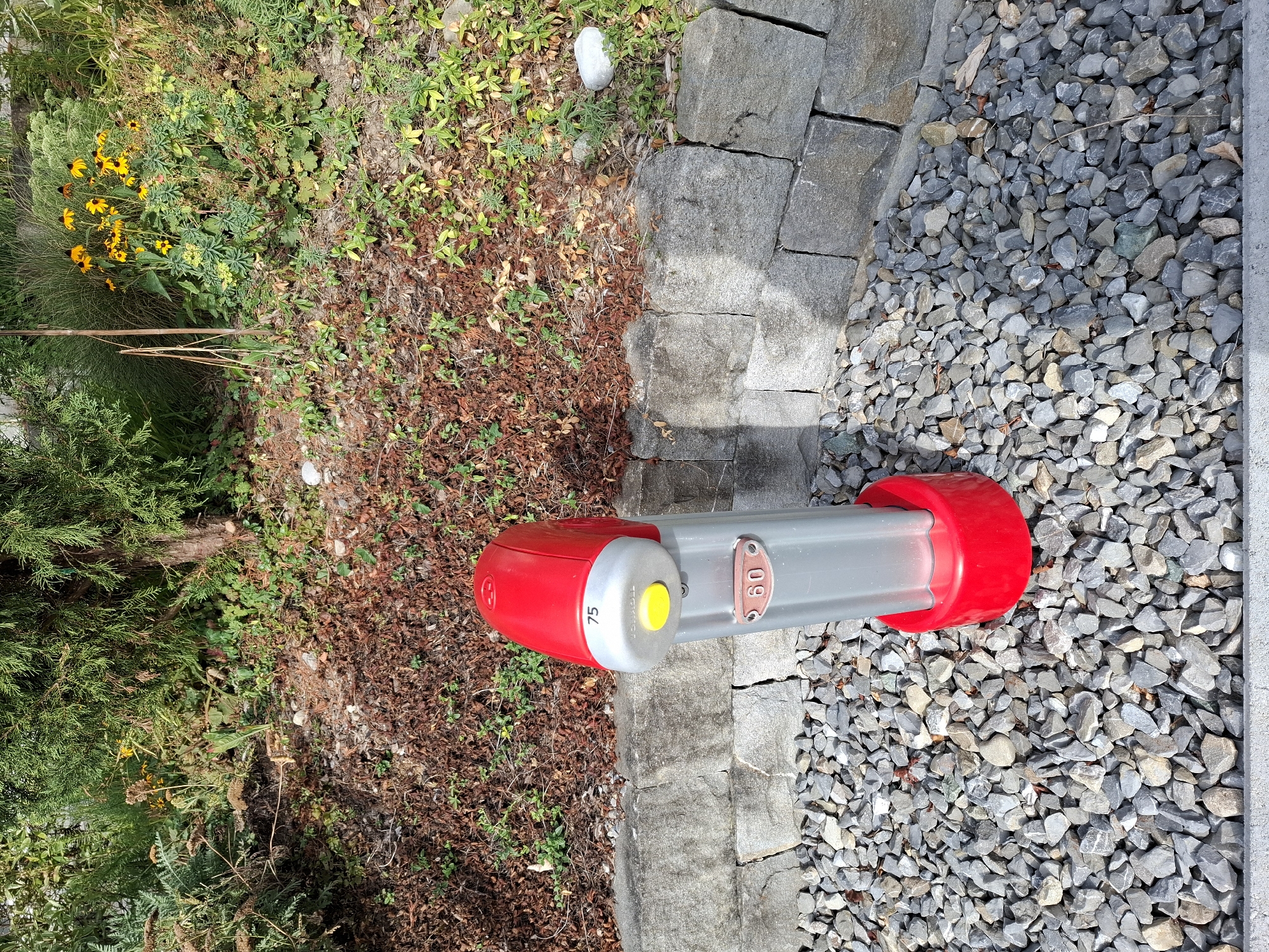 A red and silver-colored multi-functional bollard is situated on a bed of grey gravel in front of a stacked stone wall. The bollard features a yellow button, a label with the number "75", and another label with the numbers "60" inside an oval. Behind the bollard and stone wall, there is a slope covered with mulch and greenery, including flowering yellow plants. The scene appears to be outdoors in a naturalistic setting, likely a park or garden area, with a mature evergreen tree providing shade from the left.