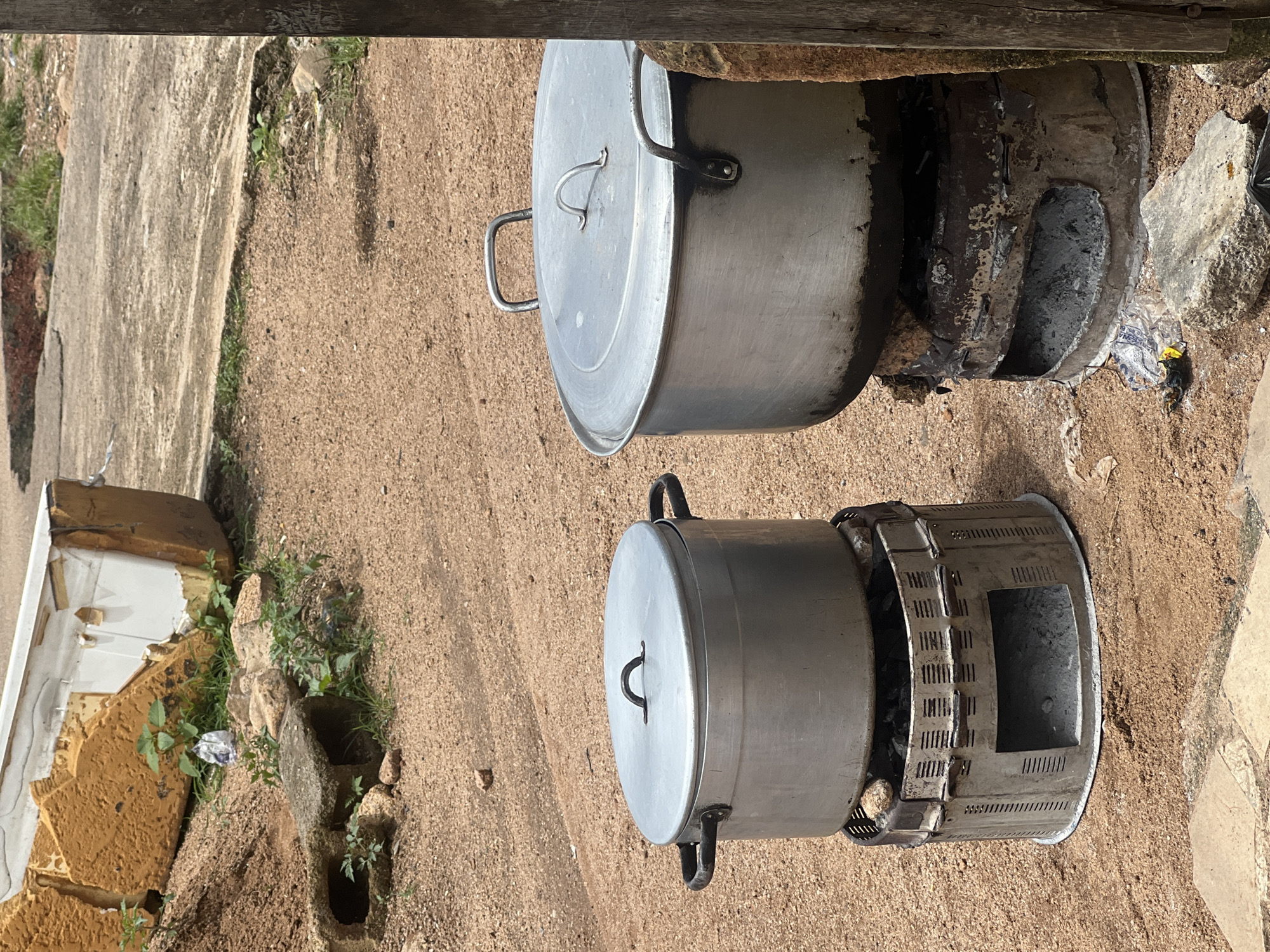 Two large aluminum cooking pots with lids are placed on metal charcoal stoves in an outdoor environment. The ground is covered in sandy soil with scattered rocks and some patches of green vegetation. In the background, there is debris including what appears to be parts of a discarded mattress or foam padding and some concrete. The overall setting suggests a rustic or informal cooking area, possibly in a less developed urban or peri-urban setting, such as might be found in the city of Jos, Nigeria, where such cooking methods are common. There are no people visible in the image.