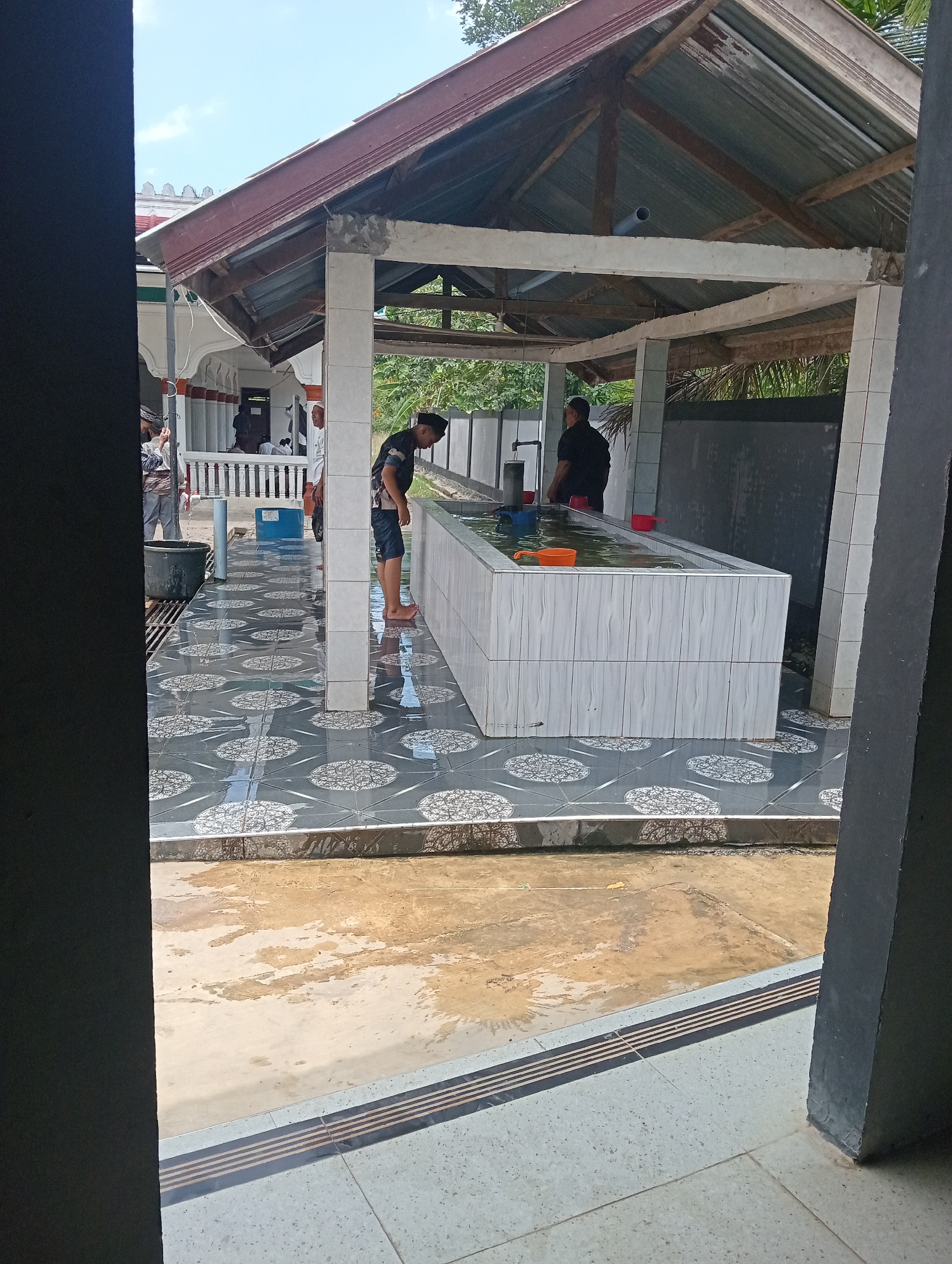The image shows an outdoor ablution area in what appears to be a mosque in Lhokseumawe, Indonesia. Two men stand at a long, rectangular ablution basin tiled in white and grey, with water inside. Smaller plastic containers float in the water. The men seem to be performing their ritual cleansing.
The ablution area is covered by a simple roof structure supported by columns, also tiled. The floor is grey tile with a circular pattern. Water is splashed on the tiles.
In the background, a building with a white facade is visible, with other men present. The foreground reveals a glimpse of an adjacent interior space, suggesting a building's doorway. The area seems to be damp, possibly from the ablutions.