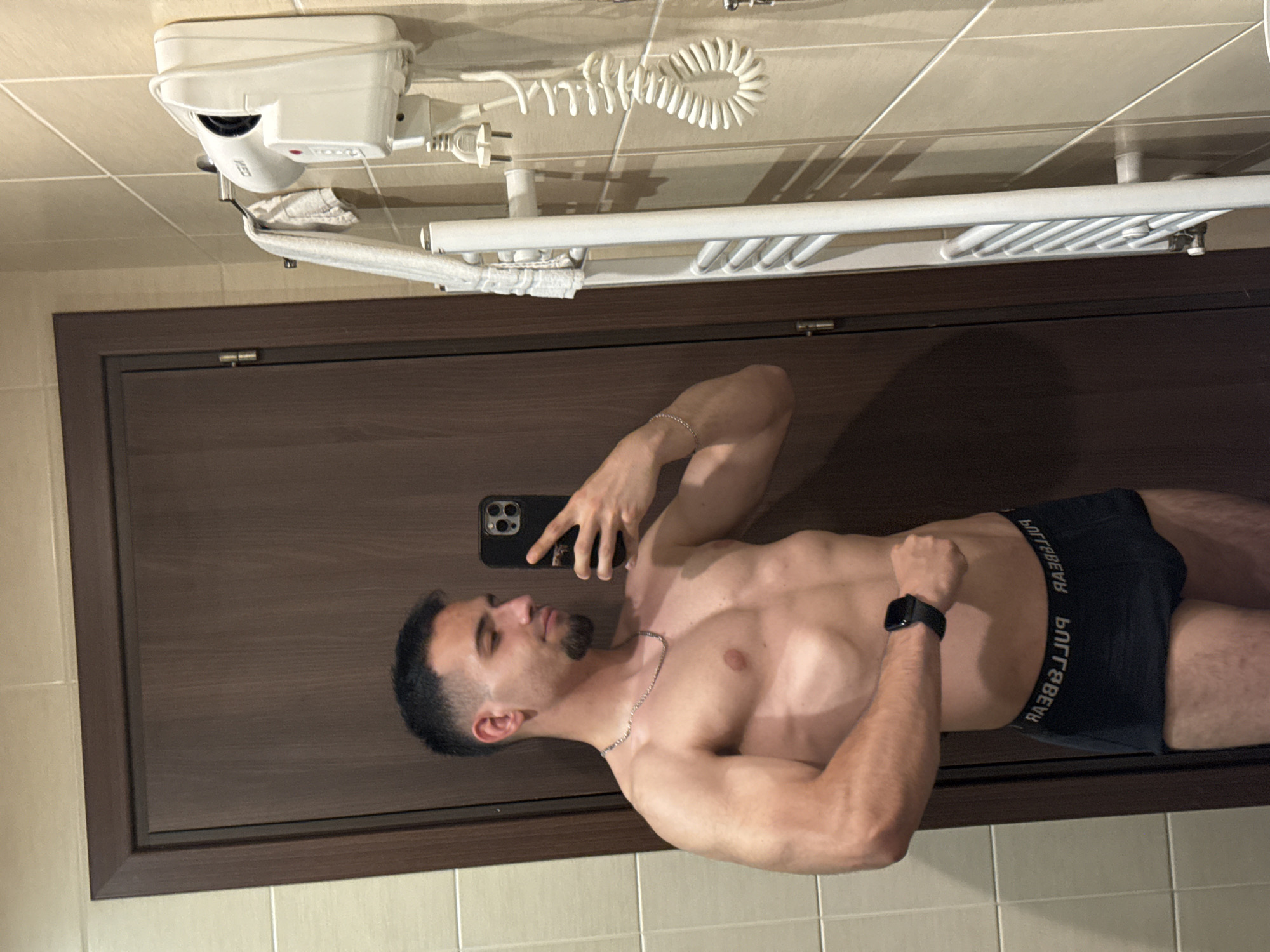 A man is posing in a bathroom. He is fair-skinned with dark hair and a short beard. He is wearing dark underwear. He holds a black smartphone in his left hand to take a picture of himself. He also wears a black smartwatch on his right wrist. The bathroom walls are tiled with light beige squares. A brown door with a dark frame is visible behind him. Above him is a white electric hair dryer and a white heated towel rack. The image was not taken in Veliko Tarnovo, Bulgaria.