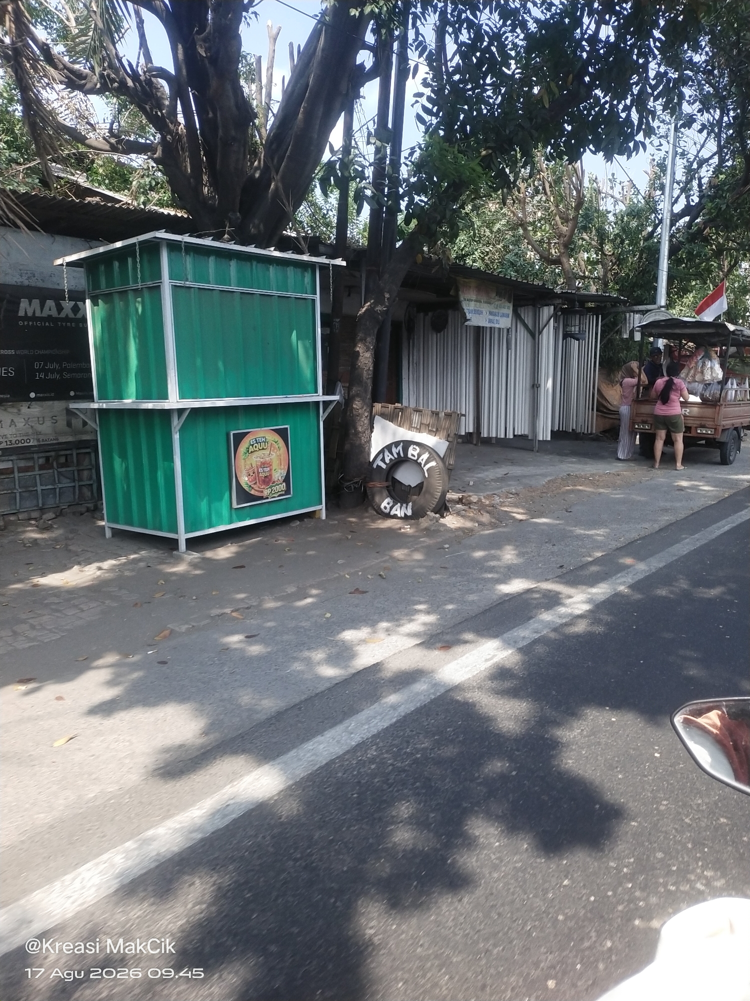The image captures a street scene in Surabaya, Indonesia. In the foreground, a paved road is visible with white lane markings and shadows cast by trees and overhead structures. A portion of a motorcycle's side mirror and handlebar is reflected in the lower right corner.
To the left, a green, modular stall stands on the sidewalk. It appears to be a food or drink vendor, with a sign on its front advertising "ES TEH AQU" and a price of "RP 2000". Above this stall, a larger structure, possibly another part of the stall or a roof, is visible, along with a large tree with bare branches.
Further back, a row of stalls with corrugated metal siding lines the street. A small, three-wheeled vehicle, resembling a delivery cart, is parked in front of these stalls. Two women are standing near the cart. One, wearing a pink top and green shorts, faces away from the camera. Another, in a pink top and striped trousers, is partially obscured. A red and white Indonesian flag is attached to a pole behind the cart.
Adjacent to the green stall and in front of the metal-sided stalls, a discarded tire with the words "TAMBAL BAN" (tire repair) painted on it lies on the ground. The overall environment suggests a commercial or market area on a sunny day. A watermark at the bottom left corner reads "@Kreasi MakCik 17 Agu 2026 09.45".