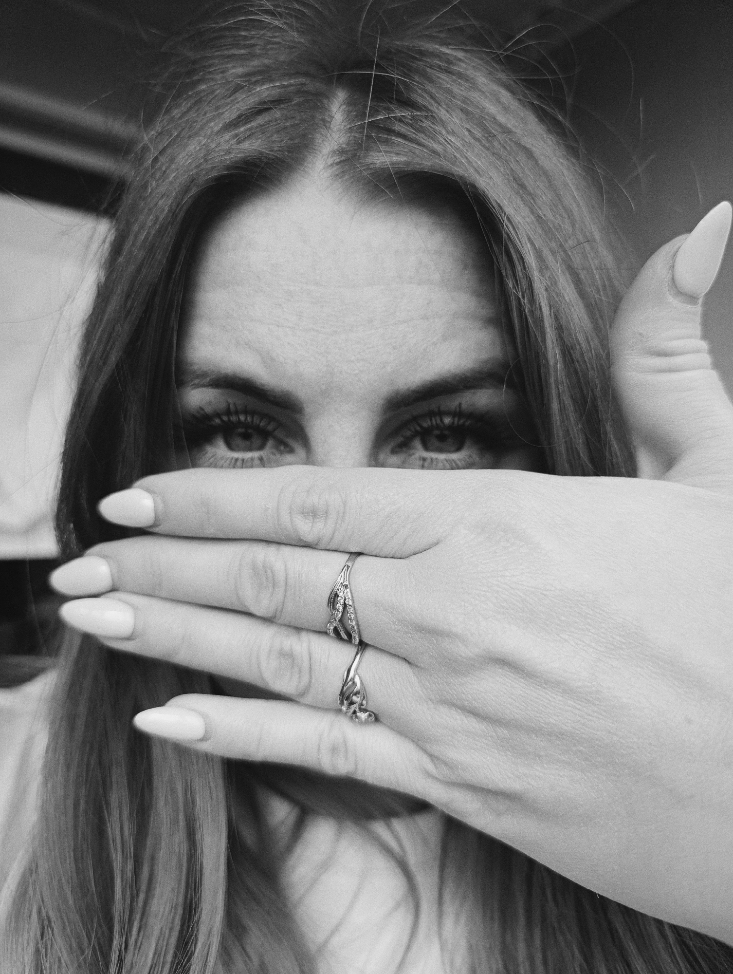 A black and white, close-up portrait captures an individual, likely female, covering the lower part of their face with their right hand. The subject's long, dark hair, parted in the middle, frames their face, with some strands visible near the temples and flowing downwards. Their eyes, enhanced with apparent eye makeup including defined lashes and brows, are wide open and gaze directly at the viewer with an intense and direct expression.
The right hand is positioned horizontally across the face, obscuring the nose and mouth. The nails are notably long, manicured, and appear to be almond or stiletto-shaped, painted with a light polish. Two rings are visible on the fingers of this hand: one on the ring finger features a detailed design with multiple small, reflective elements, and another on the middle finger has a simpler, possibly intertwined or infinity-style pattern. The skin on the hand shows subtle texture.
The background is dark and softly blurred, offering no specific location cues and ensuring the subject remains the sole focus of the image. The lighting highlights the contours of the face and hand. The gesture of partial concealment combined with direct eye contact creates an enigmatic and engaging mood. There is no visible text in the image. The black and white tonality lends a classic and artistic quality to the photograph.