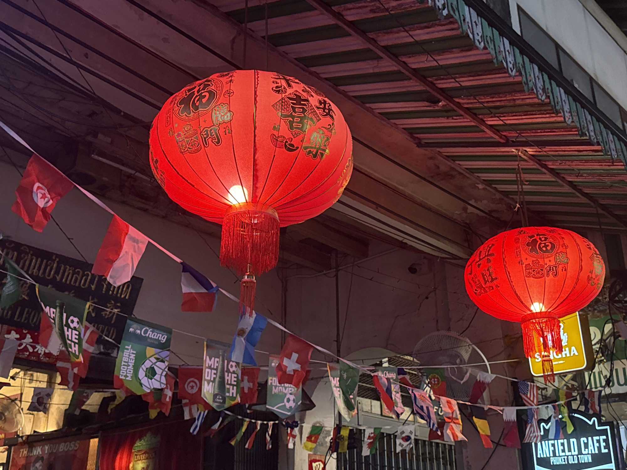 The image depicts a street scene in Phuket City Municipality, Thailand, characterized by a vibrant and festive atmosphere. Two large, red Chinese lanterns, illuminated from within, hang prominently in the foreground, adding a warm glow to the scene. These lanterns are adorned with gold Chinese characters, suggesting a celebration or cultural event.
Below the lanterns, a string of diverse national flags and banners is suspended across the street, creating a colorful canopy. These flags include those of Tunisia, Argentina, Switzerland, Portugal, and others, alongside promotional banners for "Chang" beer and "Football Moment." The presence of numerous flags and football-themed banners suggests a sports-related event or a venue celebrating international football.
In the background, the architecture appears to be typical of a bustling urban street in Southeast Asia. A red-tiled roof structure is visible overhead, supported by wooden beams. Electrical wires and conduits are strung across the ceiling, indicating an active and populated area.
Further details include a ceiling fan mounted on the wall and signage for "ANFIELD CAFE" with the subtitle "PHUKET OLD TOWN," clearly indicating the location. A smaller sign reads "THANK YOU BOSS." The overall impression is a lively street scene, possibly a market or entertainment district, decorated for a festive occasion, with a strong emphasis on international sports and local businesses.