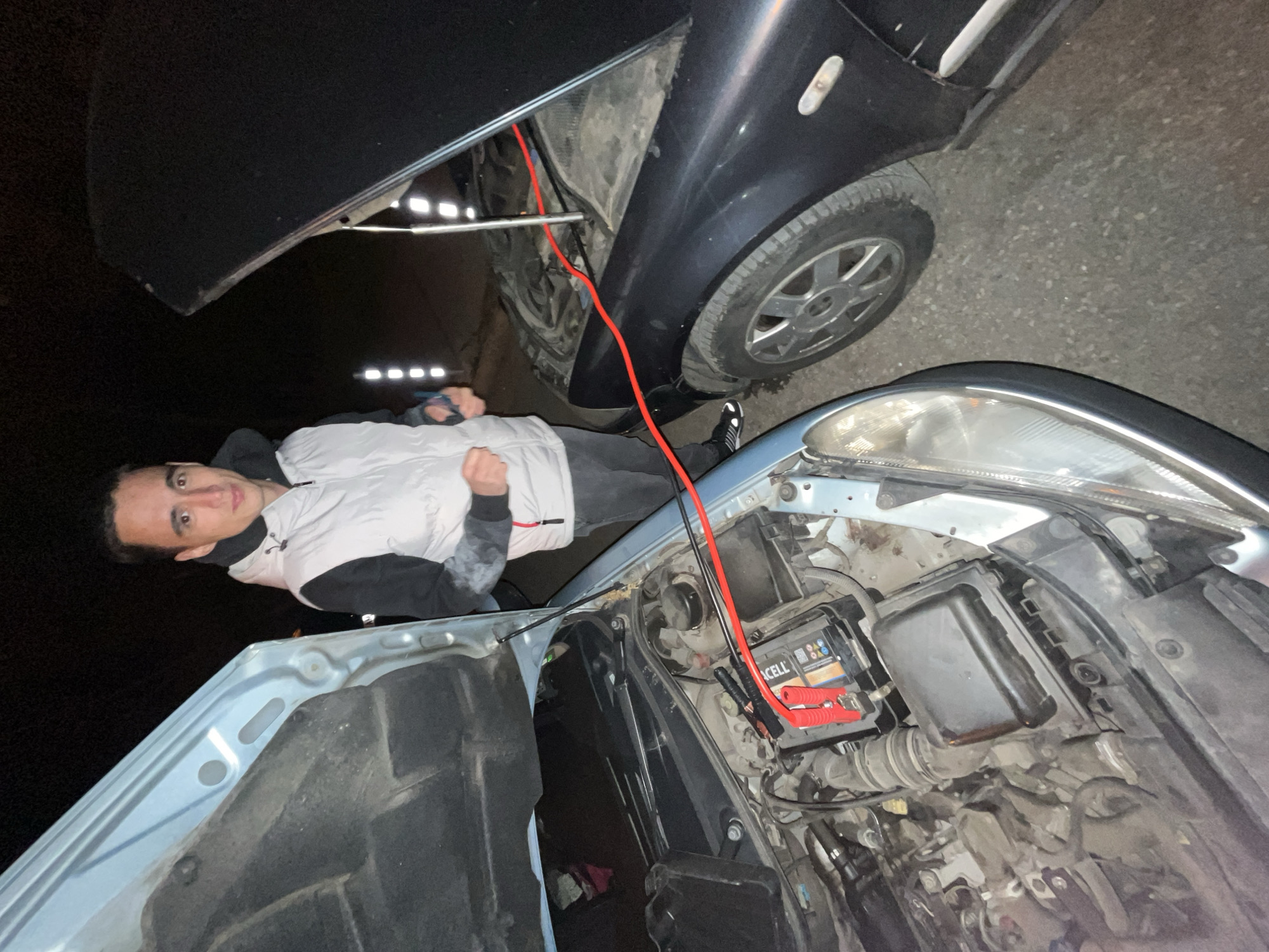A man is attempting to jump-start a light blue vehicle using a black vehicle. The hoods of both cars are open, and jumper cables are connected to the batteries. The scene appears to be on a road at night. The location of the scene cannot be determined from the image.