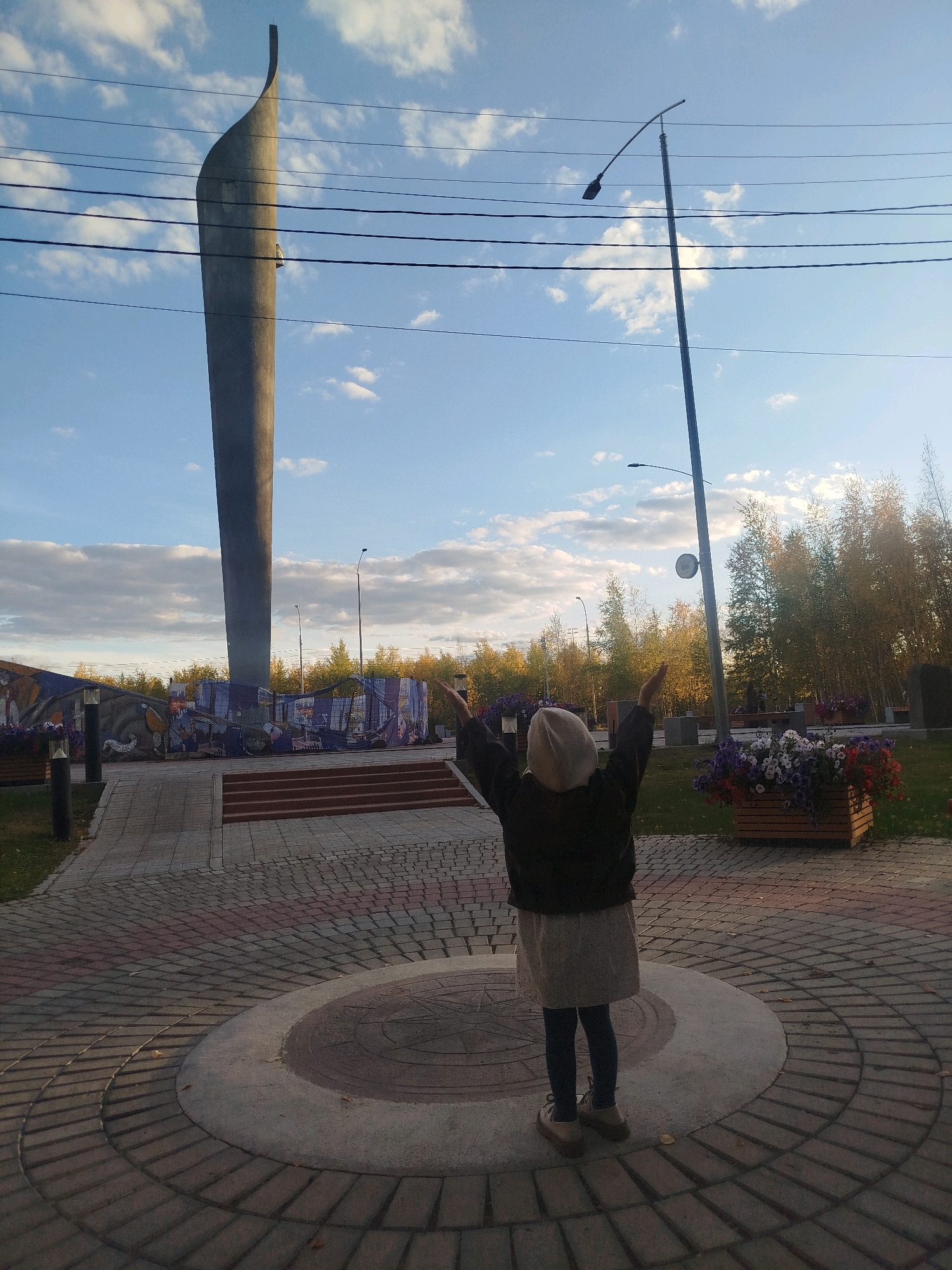 The image shows a young child, viewed from behind, standing with arms raised towards a tall, grey, abstract monument. The child is wearing a light-colored hat, a dark jacket, and a light-colored skirt with dark leggings and shoes. The monument is a large, curved, tapering structure that dominates the background. In front of the monument is a mosaic or mural depicting various scenes. The child stands on a circular paved area with a compass rose design at its center. To the right of the child are planters filled with colorful flowers. Behind the monument, trees with autumn foliage are visible under a blue sky with scattered clouds. A streetlamp stands to the right of the monument, and power lines stretch across the sky. The scene appears to be an outdoor public space, possibly a memorial or park in the city of Mirny, Russia, given the context.