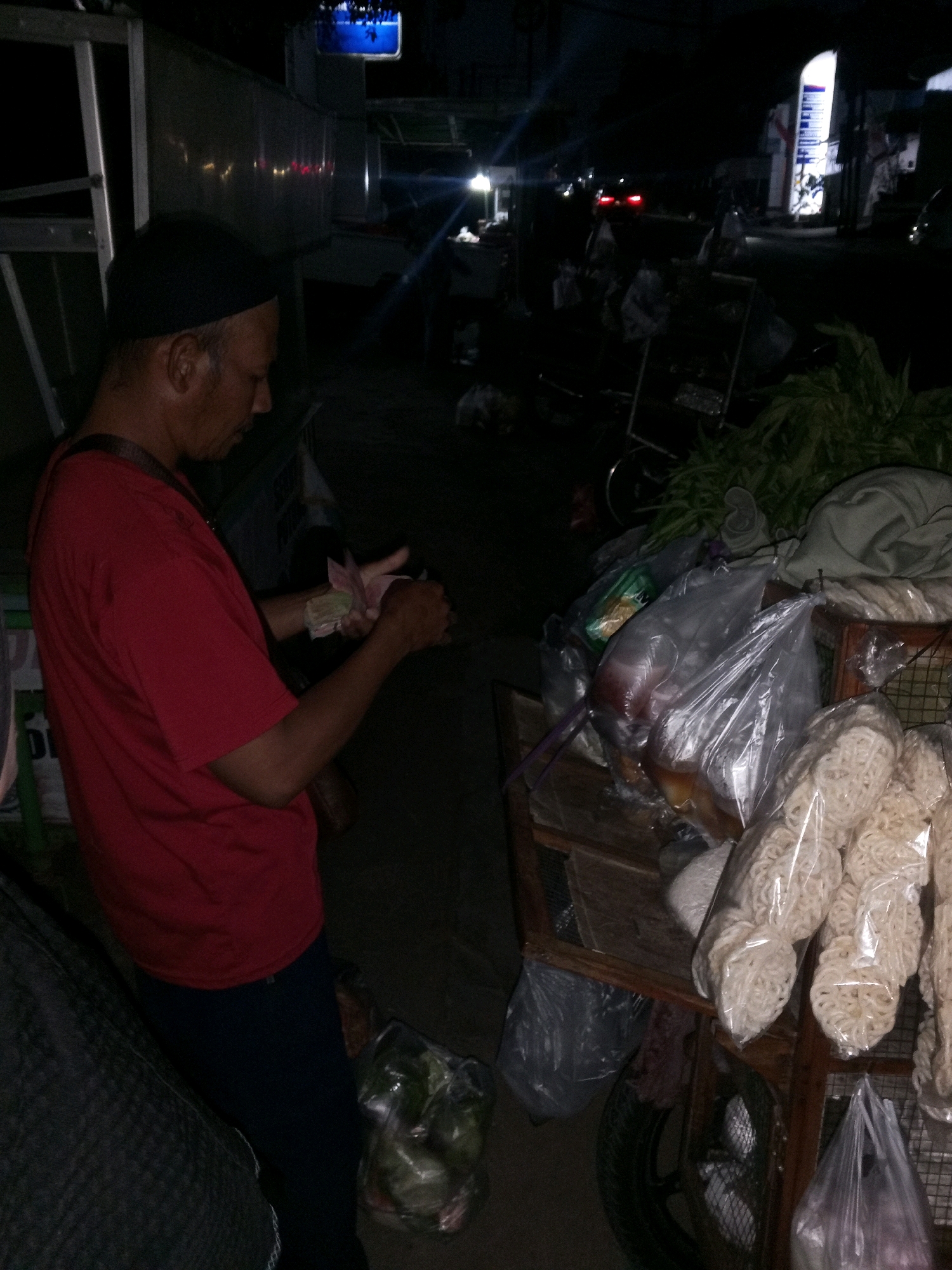 In Gentan, Indonesia, at night, a man wearing a black cap and a red t-shirt is seen standing next to a street vendor's cart. He is holding a stack of Indonesian Rupiah bills and appears to be counting or organizing them. The cart is laden with various items, including bags of what appear to be crackers or puffed snacks, some in transparent plastic packaging. There are also other bagged items on the cart, one of which contains what looks like fresh produce. The background is dark, suggesting a street scene at night, with some faint lights and reflections indicating activity or structures in the distance. The overall impression is that of a nocturnal transaction or preparation at a local street market or roadside stall.