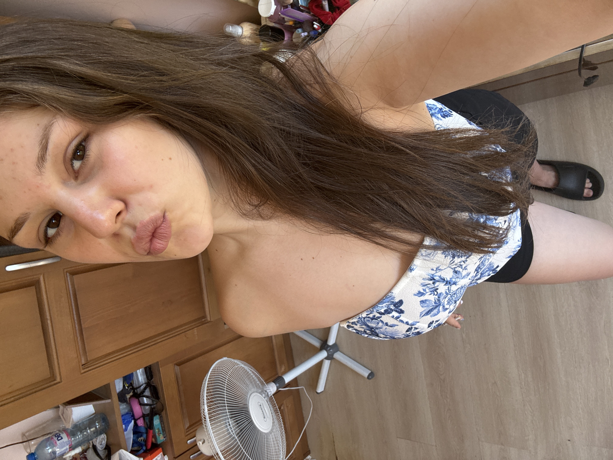 A young woman with long, brown hair is taking a selfie. She is wearing a blue and white floral-patterned top and black shorts. She is standing in front of a wooden cabinet, with a fan to her left. The floor is made of light-colored wood. In the background, shelves are visible, stocked with various items, including a water bottle and toiletries. The woman is looking at the camera with her lips pursed.