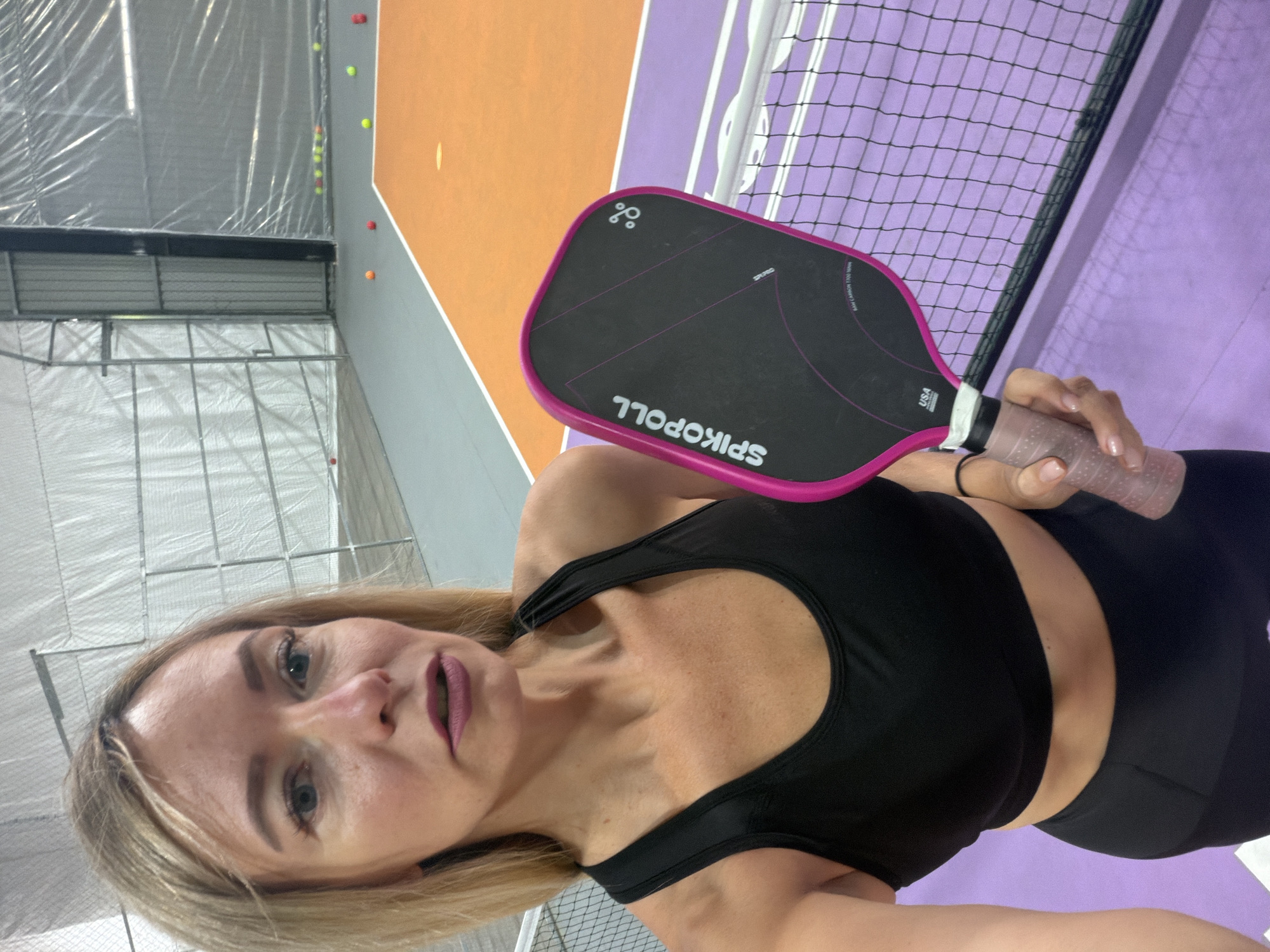 An athletic woman with blonde hair is taking a selfie in an indoor pickleball court. She is wearing a black sports bra and black leggings. She is holding a pink and black pickleball paddle. The court has a purple and orange surface with a net in the foreground. The background consists of white netting and metal poles. There are several pickleballs scattered on the court. The scene appears to be indoors, possibly a sports facility. It is not possible to determine the city of Da Lat, Vietnam from the provided image.