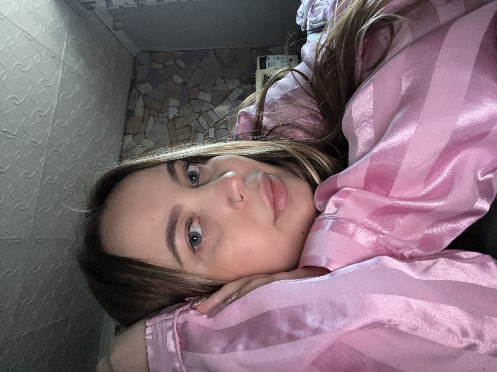 A woman is lying on her side, resting her head on her hands, which are positioned on top of a pink striped satin shirt or pajamas. She has light brown hair with blonde highlights and fair skin. She is wearing a nose ring. The wall behind her features a mosaic pattern with light brown and grey stones. To the left of the mosaic wall is a textured white wall. The location is unknown.