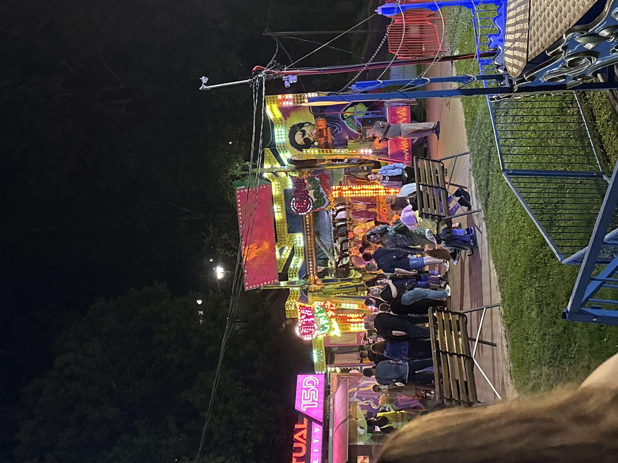 The image shows a busy amusement park scene at night, with a large, brightly lit ride in the center. The ride is colorful and features flashing lights, with various people gathered around it. Some are standing and watching, while others are sitting on benches. There's a sign in neon pink that says "TUAL 15D" on the left side of the frame. Another sign on the ride says "Holy Bobs" in colorful lettering. A large graphic of a man wearing sunglasses is displayed on the ride. The ground is paved in some areas and covered with grass in others. Trees are visible in the background, illuminated by the park's lights. The overall atmosphere suggests a lively event. This scene is located in the city of Vitsebsk, Belarus.