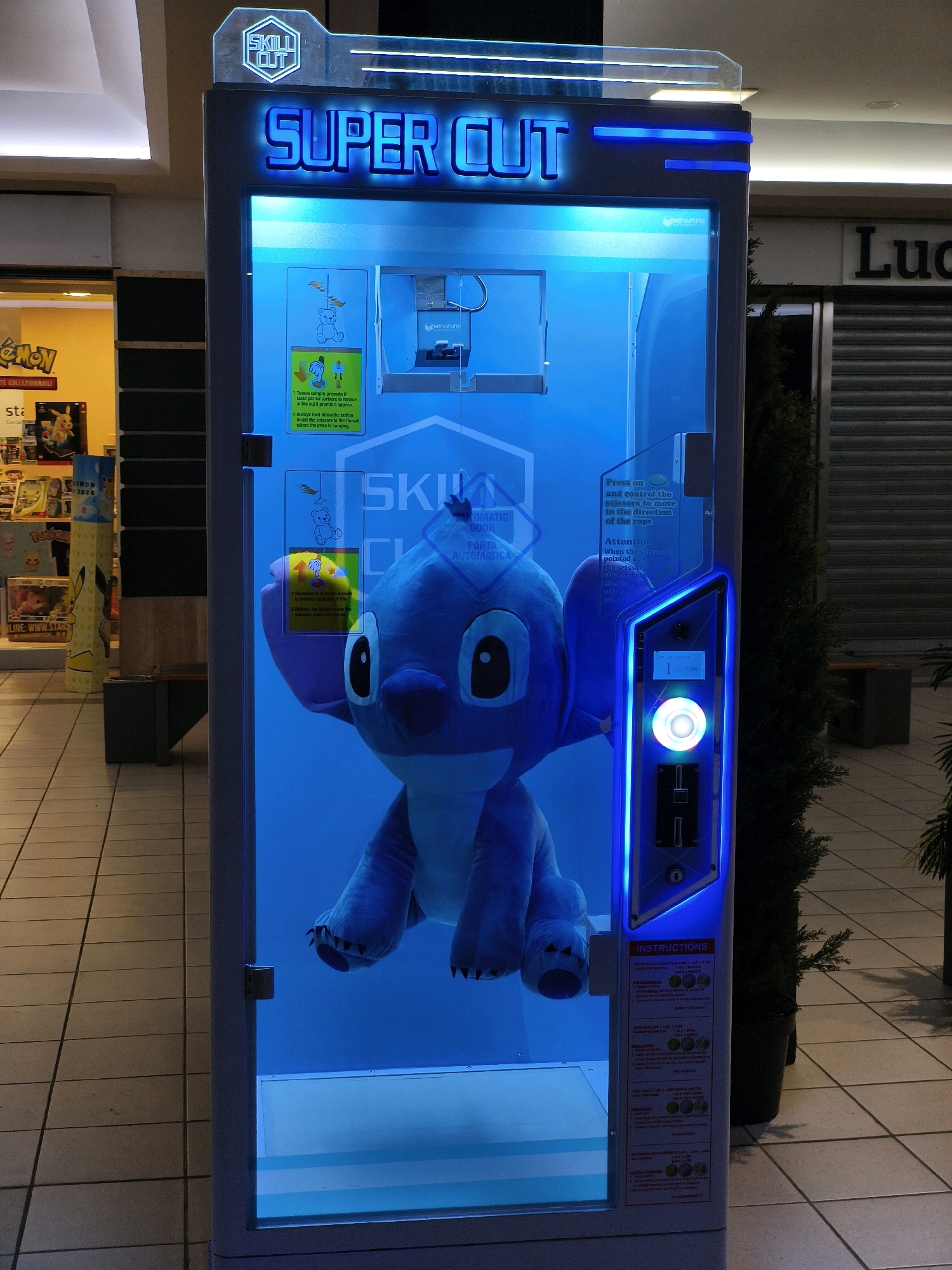 The image shows a close-up of a "SUPER CUT" claw machine, which contains a large plush toy of the character Stitch from Disney's Lilo & Stitch. The machine is illuminated with bright blue LED lights. Text on the machine, some in Italian, indicates it is a skill game with instructions for operation. To the left of the machine, a retail store is visible with displays of Pokemon merchandise. The flooring is tiled. The overall environment suggests an indoor public space, likely a shopping mall or arcade. The city of Chiugiana, Italy is not discernable from the image.