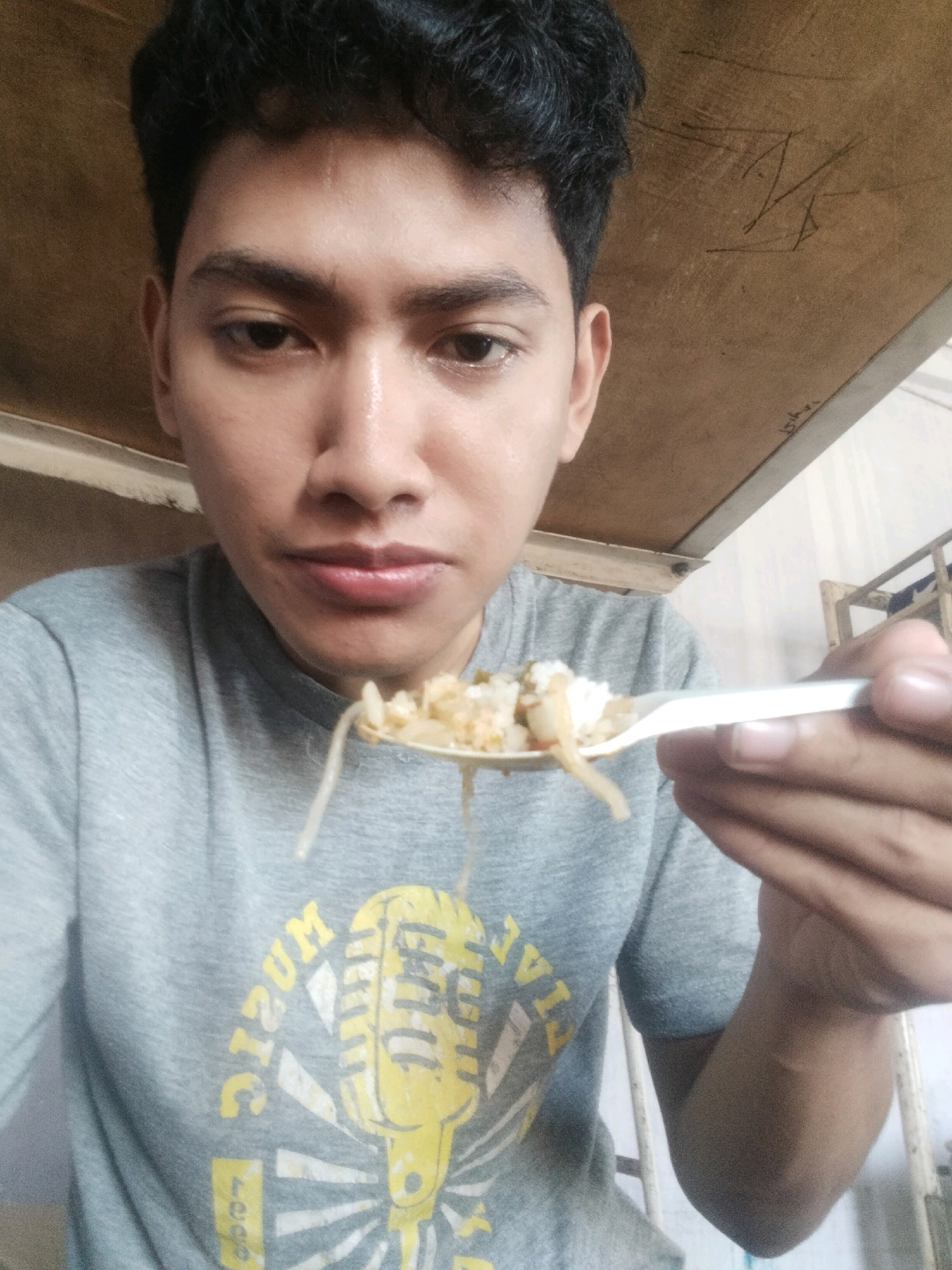 A young man is eating from a spoon in this close-up selfie, holding the spoon with his right hand. He is wearing a grey t-shirt with a yellow graphic of a microphone and the word "MUSIC" on it. The background appears to be a wooden ceiling or shelf above him, with some faint markings. To his right, a blurry white structure, possibly part of a bunk bed or shelving unit, is visible. The lighting suggests an indoor setting. The man's facial features are in focus, showing his eyes, nose, and lips as he prepares to eat. The image is taken from a frontal perspective. The location is not specified within the image itself but the query states City of Medan, Indonesia.