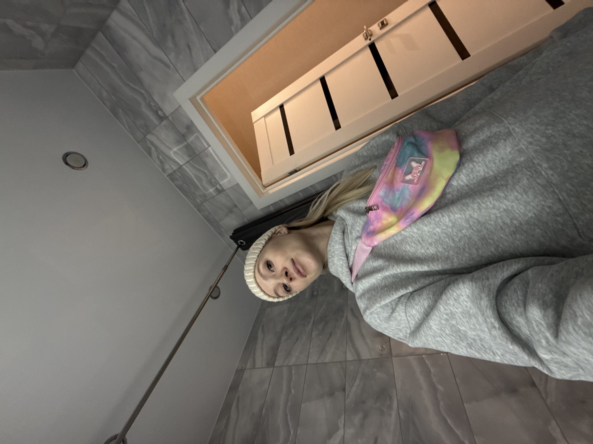 A woman with blonde hair and wearing a white beanie is standing in a bathroom. She is wearing a grey hooded sweatshirt and a tie-dye fanny pack. The bathroom has light grey marble-patterned tiles on the walls and floor. There is a shower rod on the left side of the image. A white cabinet with black accents is above and behind the woman. The location is an interior space, presumably a bathroom. There is no contextual information to suggest a location in Izhevsk, Russia.