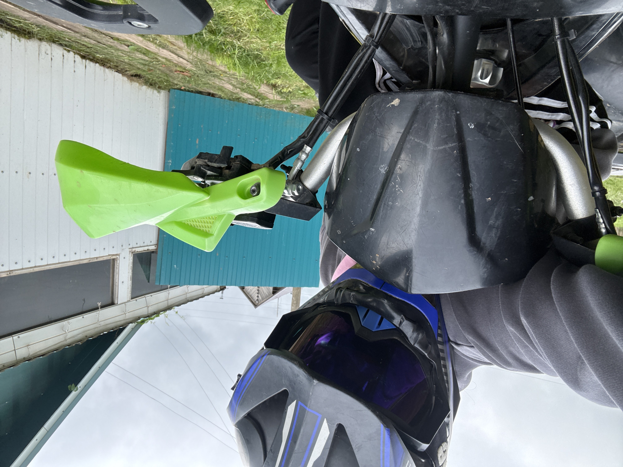 The image captures a first-person perspective from a person seated on an off-road vehicle, likely an ATV, in Ляльшур, Russia. The individual wears a black and blue full-face helmet with a dark, reflective visor, a grey hooded sweatshirt, and black pants. The vehicle's handlebars are silver, with a scratched black plastic fairing positioned centrally. A bright green plastic handguard is visible on the right handlebar. Cables and hoses, secured with black zip ties, are evident around the steering column. The ground in the immediate vicinity is covered with green grass and dirt. The background features an overcast sky above a teal corrugated metal fence or gate. Behind this fence is a white building with dark rectangular windows and a dark green pitched roof. Some vegetation is visible along the roofline. Utility wires are present against the cloudy sky, and a portion of a wooden building's roof is discernible in the far background.
