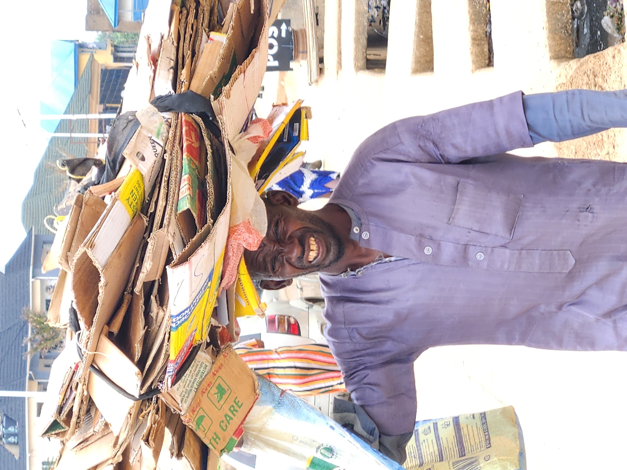 This vibrant image, likely captured in Jos, Nigeria, features a man expertly balancing a colossal stack of flattened cardboard boxes on his head. The man, appearing to be of African descent, has a beard and is dressed in a light purple/grey long-sleeved shirt, possibly a traditional garment. He is smiling broadly, revealing a joyful expression and teeth, which conveys warmth and friendliness.
The towering load of recyclables consists primarily of diverse cardboard pieces, secured tightly with dark bands, showcasing an impressive feat of balance and strength. Visible text on some of the boxes includes "TH CARE," "K," "DISTRIBUTED BY Euro Mega Atlantic Nigeria Ltd. 1. Henry Carr Street, Off Ak," "600," "126G INSTANT," "STY," "EN," and "QUEEN." A faint sign with "POS" and an upward arrow can also be discerned in the background.
The setting appears to be an outdoor urban or semi-urban environment, with various buildings, some featuring blue roofs, and light-colored concrete structures in the background. The bright, clear lighting indicates it is daytime, likely a sunny day. The man's activity of transporting collected materials on his head is a common sight in many regions, emphasizing resourcefulness and physical endurance. His beaming smile adds a notable emotional dimension to the scene.