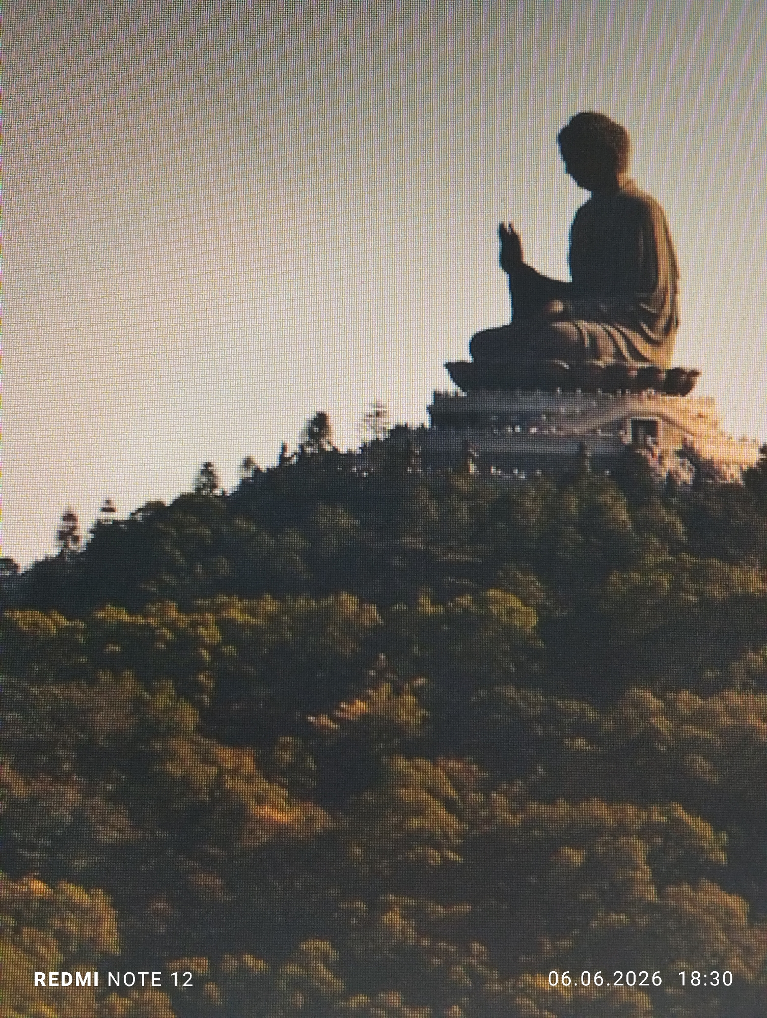 The image displays a large, dark silhouette of a seated Buddha statue, prominently positioned on a multi-tiered platform atop a densely forested hill. The Buddha's right hand is raised in a gesture, with the fingers pointing upwards. The scene is set against a bright, pale sky, suggesting either early morning or late afternoon light. The lower portion of the image is dominated by the dark green foliage of the hillside. This scene is located in the city of Burgas, Bulgaria. The image itself exhibits a visible grid pattern across its entire surface, indicating it is a photograph of a digital screen rather than a direct photograph of the scene. Overlays in the bottom left corner read "REDMI NOTE 12," and in the bottom right corner, "06.06.2026 18:30."