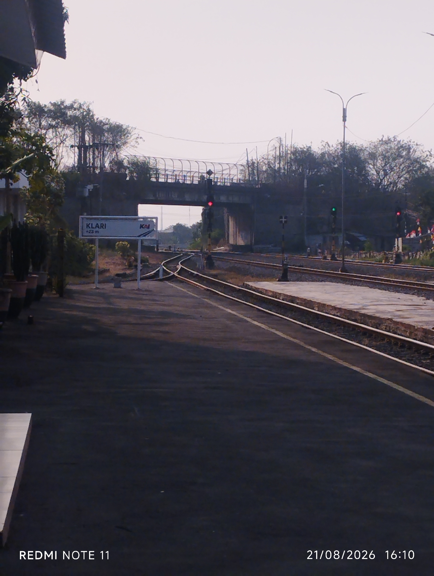 The image depicts the Klari train station, located in Karawang, Indonesia. The scene captures a perspective of the train tracks at the station, with a platform on the left and a bridge overhead. A sign indicating "KLARI +23 m" and the KAI logo is visible. The sky is overcast, and the lighting suggests it is either early morning or late afternoon. There are no people clearly visible in the immediate foreground or on the platform, though figures can be faintly seen in the distance near the bridge. The tracks are well-maintained, and there are no trains present in the shot. The environment appears to be an active but not busy train station.