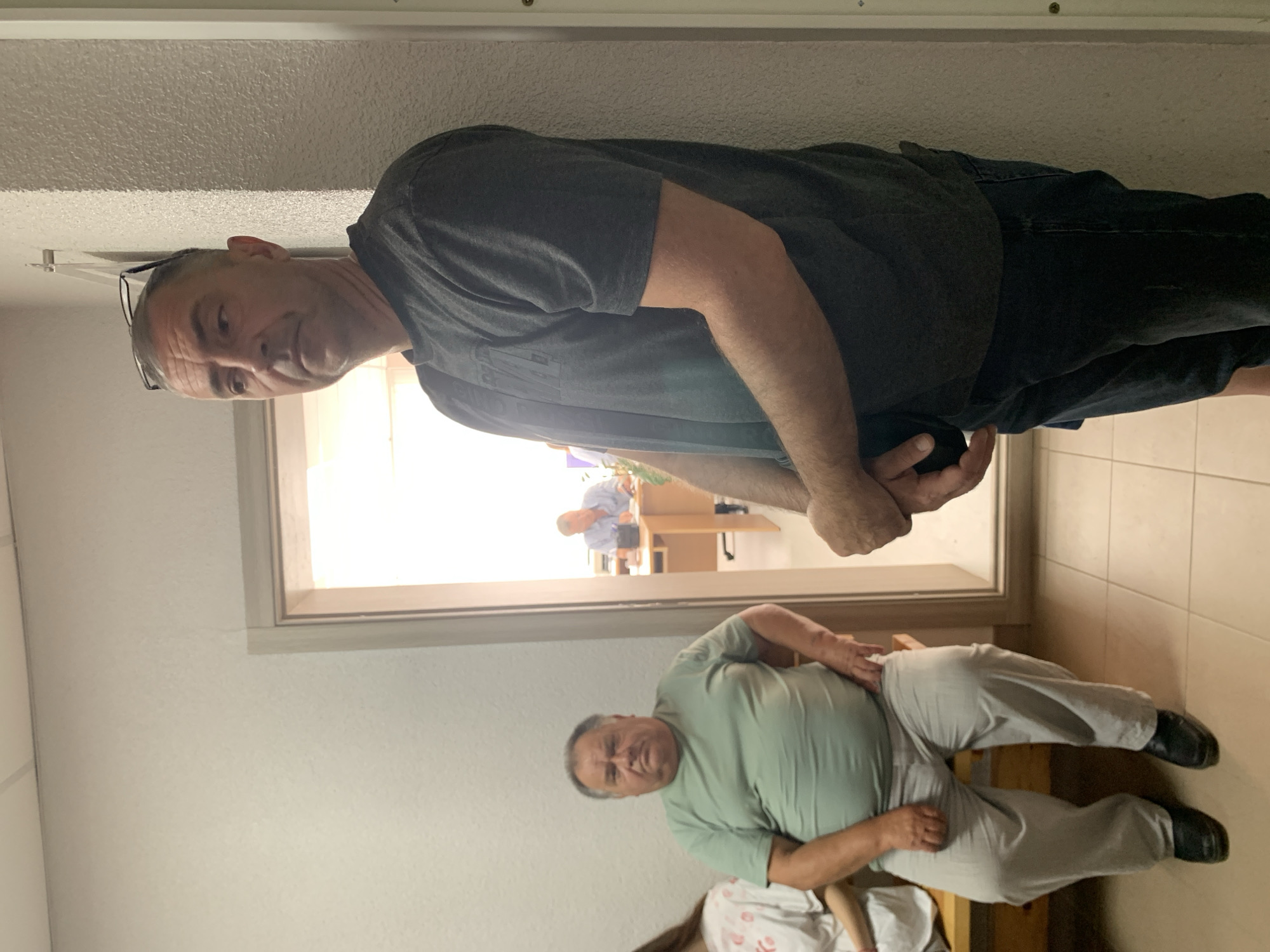 The image depicts an indoor scene, possibly a waiting area or hallway, with a light-colored tiled floor and off-white walls. The overall lighting suggests an indoor environment, possibly during the day.
In the foreground, a middle-aged man with short gray hair and glasses perched on his head is standing, leaning against a textured off-white wall. He is wearing a dark gray t-shirt and dark pants. His gaze is directed to his left, away from the camera.
To the left of the standing man, a doorway is visible, leading to another room. Through the doorway, a man in a light blue collared shirt is seated at a desk, appearing to be engaged in work. The room beyond the doorway is brightly lit.
Further to the left, a man is seated on a wooden bench. He is wearing a light green t-shirt and light gray pants, with black shoes. He is facing forward, looking directly at the camera with a neutral expression. To his left, partially visible, is another person wearing a white t-shirt with a pink design.
The scene appears to be in a public or institutional setting within the Бокшыцкі сельскі Савет in Belarus, given the context of the request.