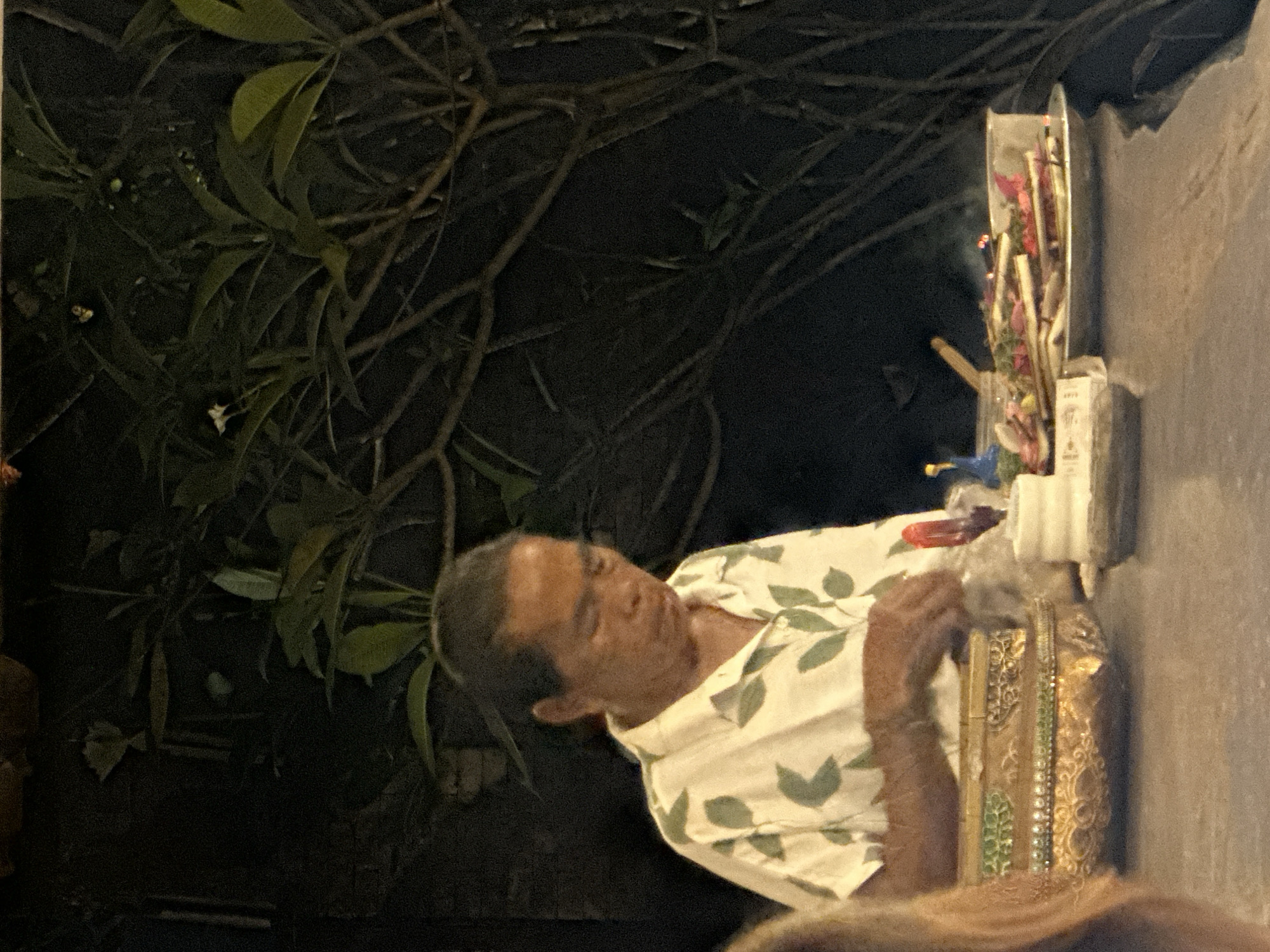 The image shows a man wearing a light-colored shirt with a green leaf pattern. He appears to be in the process of arranging or interacting with some offerings placed in a container. The offerings include what seem to be sticks or incense, and colorful flowers, possibly pink and red. There is also a cylindrical object, perhaps a vase or a container for the offerings, and a small white object that could be a card or a label. The scene is dimly lit, suggesting it might be evening or nighttime. Behind the man, there are dark branches and leaves of a plant. The man's face is visible, and he appears to be concentrating on his task. To the right of the man and the offerings, there is a textured, greyish surface that could be a wall. The overall setting appears to be outdoors, given the presence of foliage, and the dimly lit environment with hints of man-made structures. The location context provided is North Kuta, Indonesia, which might imply a cultural or religious setting where such offerings are common. There is a small piece of text visible on a white object in front of the offerings, but it is too blurry to read.
